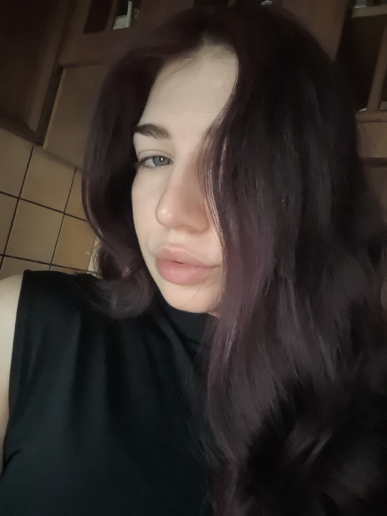 A close-up shot of a fair-skinned woman with dark plum-colored hair that cascades over one side of her face. Her hair is wavy, and a strand falls across her face. She has light blue eyes, a straight nose, and full lips. She's wearing a sleeveless black top. The background consists of what appears to be tiled walls and dark wooden cabinets, suggesting an indoor location, possibly a kitchen or bathroom. The location is in the city of Unknown, Unknown.
