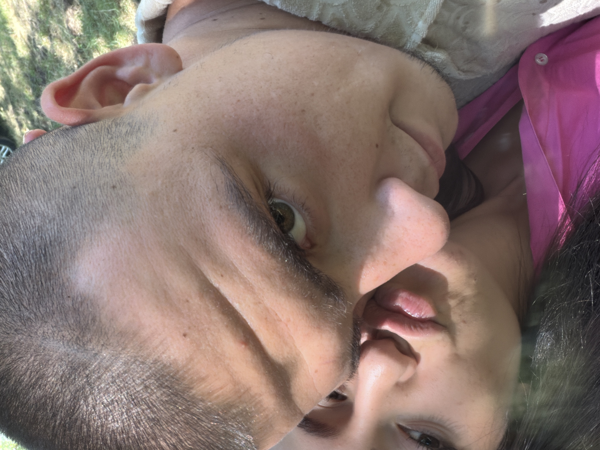 This is a close-up, selfie-style image of two people, a man and a woman, outdoors. The man is in the foreground, with his head tilted towards the camera. He has short, dark hair and is wearing a light-colored, textured garment over his shoulder. The woman is positioned behind and to the side of the man, with her face partially visible. She has dark hair and is wearing a bright pink shirt. Her lips are puckered as if for a kiss, and she is leaning her head towards the man's cheek. The background shows greenery and grass, suggesting an outdoor setting, possibly a park or garden. The overall lighting suggests it is daytime. The image does not contain any identifiable landmarks or features that would pinpoint its location in Burgas, Bulgaria.