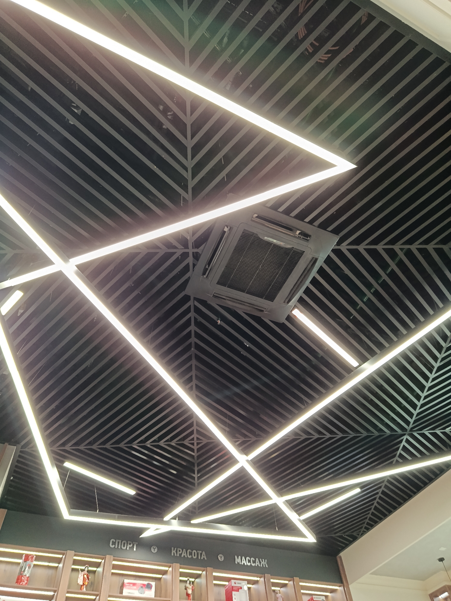 The image depicts a ceiling in what appears to be a commercial space, likely a retail store or beauty salon. The ceiling is decorated with a geometric pattern of alternating dark and light stripes, creating a visual effect of radiating lines. Integrated into this design are several long, rectangular LED light fixtures, arranged in a seemingly random yet aesthetically pleasing pattern. A square air conditioning unit is centrally mounted within the ceiling design.
Below the ceiling, at the bottom of the frame, is a glimpse of shelving displaying various products, suggesting a retail environment. The words "СПОРТ", "КРАСОТА", and "МАССАЖ" (SPORT, BEAUTY, and MASSAGE) are visible above the shelves, indicating the type of products or services offered.
Based on the signage and general aesthetic, the location may be in Moscow, Russia.