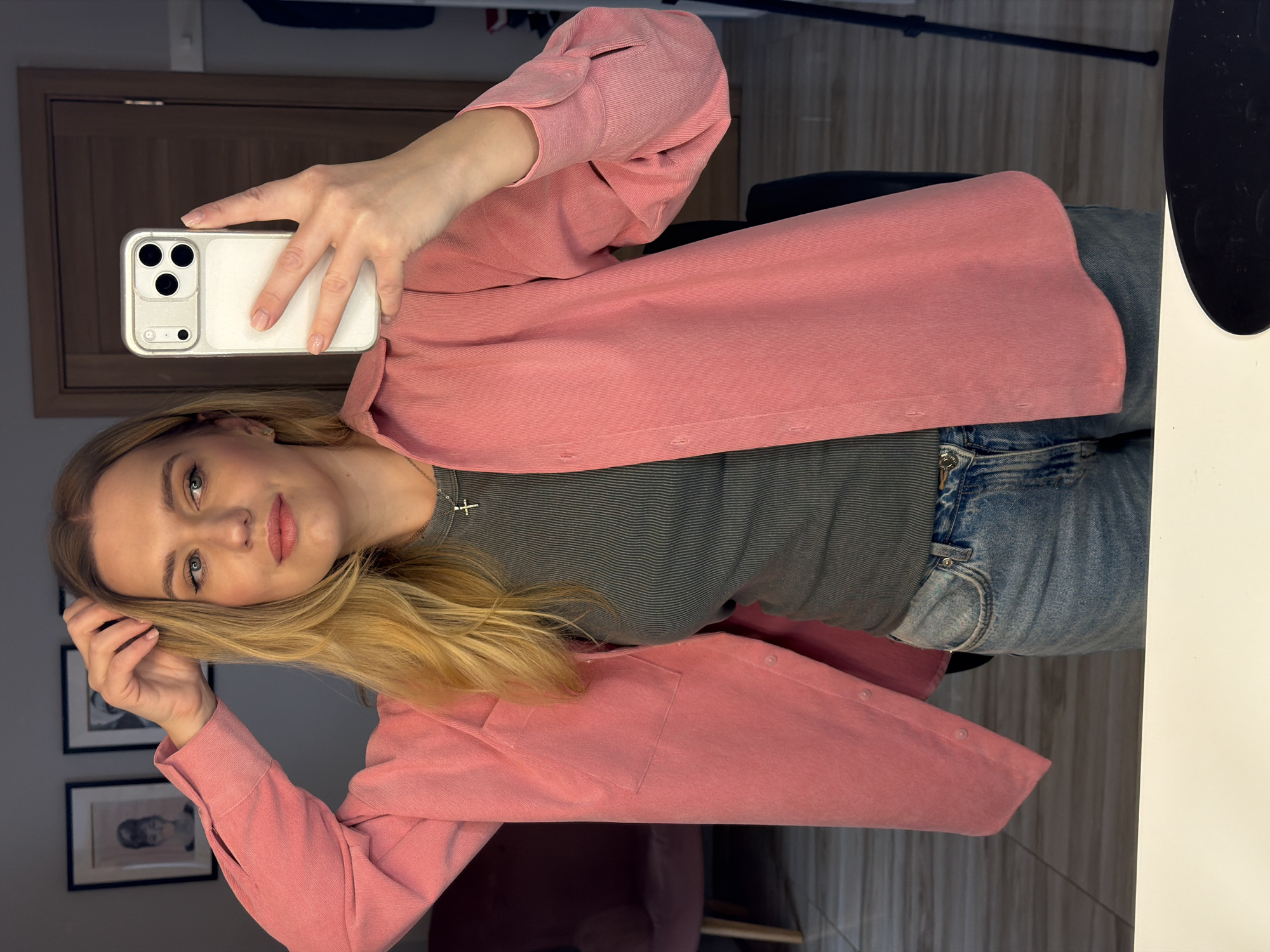 A woman is taking a selfie in an indoor setting. She is wearing a pink shirt over a striped gray top and jeans. She is holding a white iPhone in her right hand, capturing the image. Her left hand is touching her blonde hair.
In the background, there is a door, a chair, and framed pictures on the wall. The setting appears to be an interior space, possibly a home or studio. There is no specific evidence to pinpoint the location within Riga, Latvia.