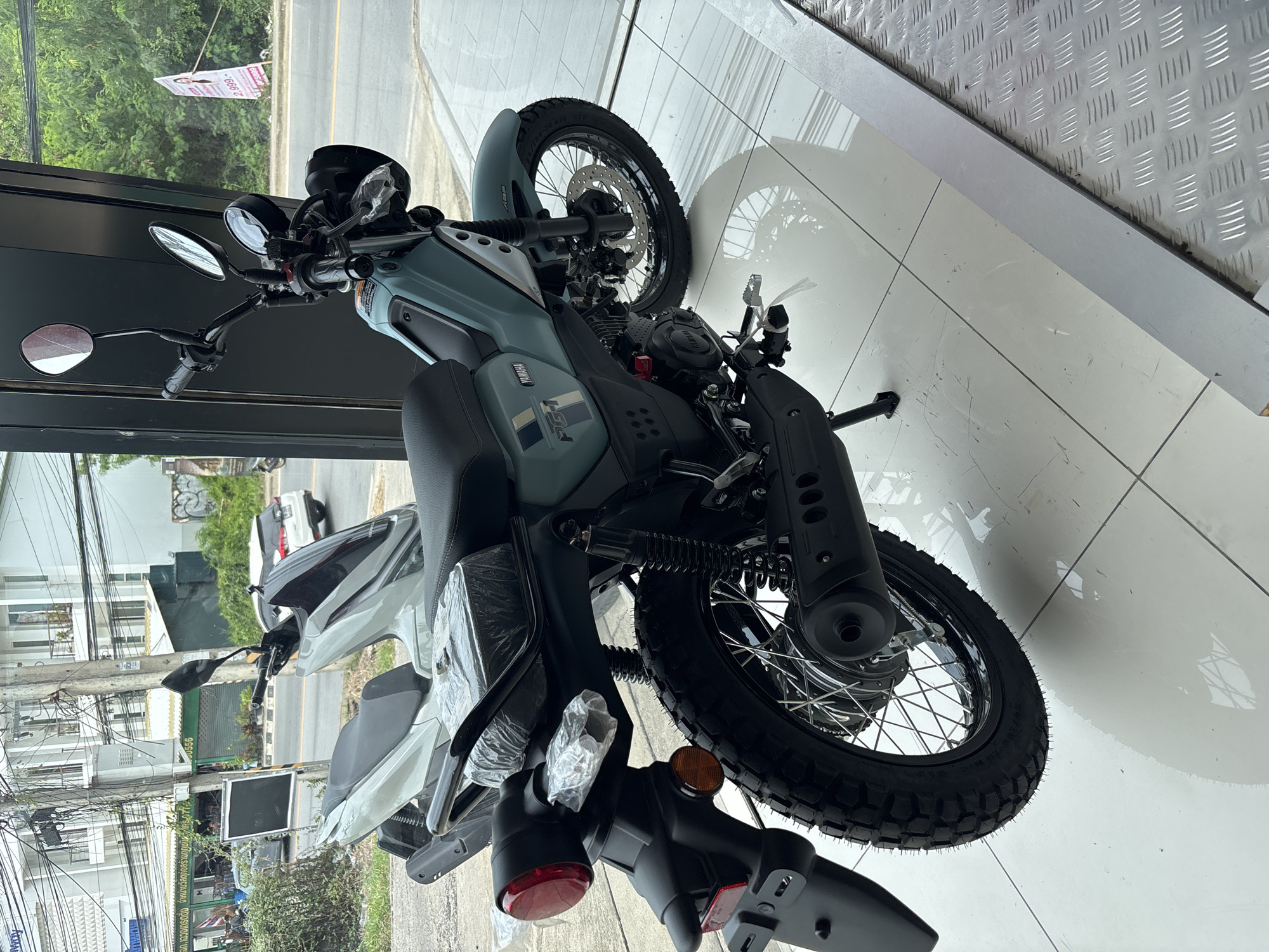 A rotated, high-angle view shows a new gray Yamaha PG-1 motorcycle in what appears to be a showroom in Baan Bo Phut, Thailand. The motorcycle is parked on a white-tiled floor. A silver, metal-textured surface is in the upper right corner.
Through the showroom window, a street scene is visible with buildings, power lines, and greenery. Another motorcycle is parked outside. A road stretches out, and in the distance, buildings and trees can be seen. A sign reads "แจก.เฉวงเทรดดิ้ง จําหน่ายวัสดุก่อส." Further down the sign, "00556" is visible. The sign also has the price "2,999.-" and "686" at the top.