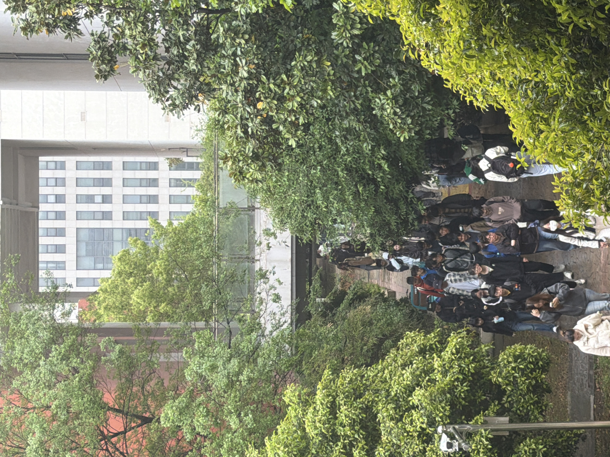 The image shows a scene in Jiangning District, China. A group of people, predominantly students, are walking along a path. The path is lined with trees and greenery on both sides. Buildings are visible in the background, including a modern building with a grid of windows. The weather appears to be overcast or rainy.