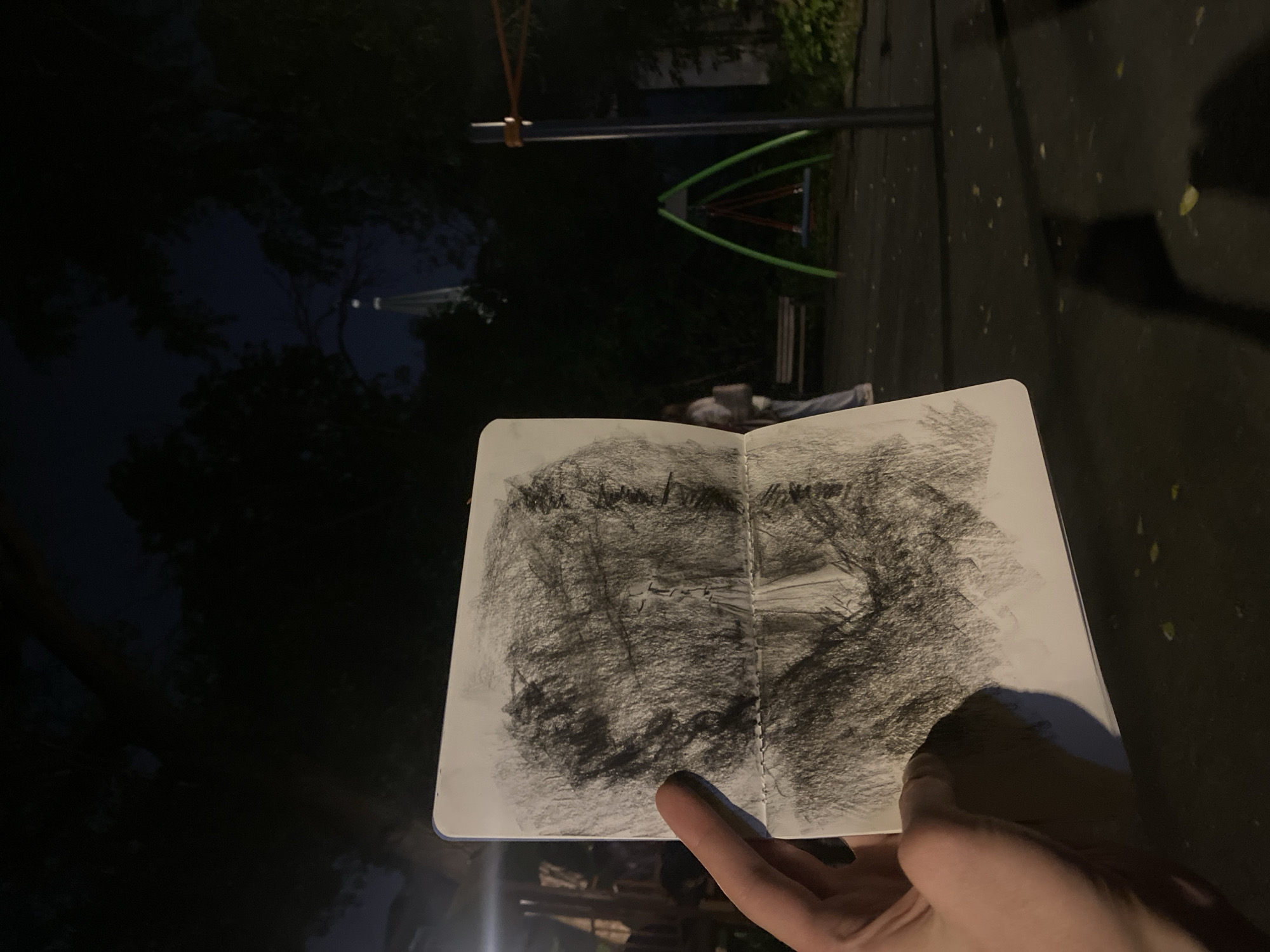 A person holds open a sketchbook in a park at night. The sketchbook displays a charcoal drawing, obscuring the view of the park. In the background, one can discern a playground structure, a bench, and a person lying down on the bench. The location is likely within the city of Moscow, Russia.