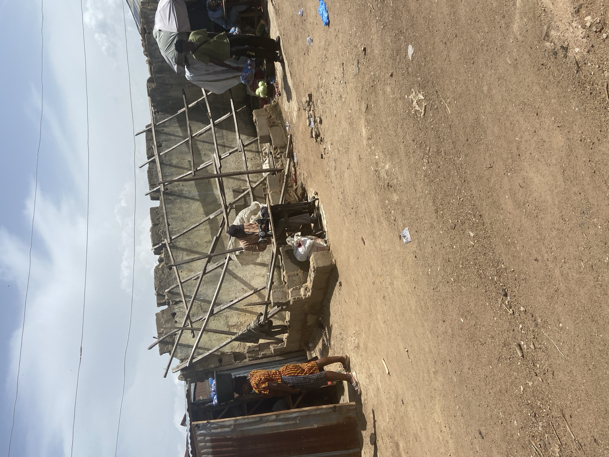 The media file shows a dusty, outdoor scene in Bukuru, Nigeria. Several people are visible, some engaged in what appears to be a market or communal gathering. In the foreground, a person wearing a brightly colored, patterned shirt and plaid shorts stands near a corrugated metal structure. Behind them, another person is seated, possibly at a table, with a laptop in front of them.
Further back, a partially constructed building is evident, with exposed scaffolding and a rough concrete wall. Several individuals are clustered near this structure. One person, wearing a light-colored garment and a head covering, is interacting with others. Another person in a green shirt is seen holding plastic bags, possibly containing goods. A white umbrella or canopy is also visible, providing shade.
The ground is a dry, dusty earth, with scattered debris, including plastic bags and what looks like small stones or rubble. The sky above is partly cloudy, suggesting it is daytime. The overall impression is one of a busy, informal setting, likely a marketplace or a community area undergoing construction. The lighting suggests it could be late morning or early afternoon. There are no legible signs or text visible in the image.