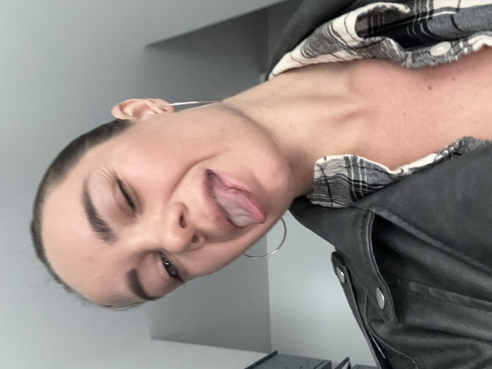 A person with fair skin is sticking their tongue out and winking at the camera. They are wearing a black leather jacket over a black and white plaid button-down shirt, and silver hoop earrings. The background is a plain, light gray wall. The image is rotated slightly. The location is not apparent from the image.