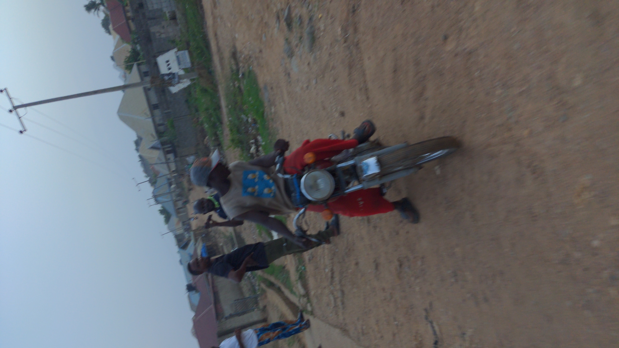 The image depicts a man riding a motorcycle on a dirt road in Ado, Nigeria. Two other individuals are standing near the road. A power pole is visible in the background, as are houses and vegetation. The rider wears red pants, a t-shirt, and a cap. The scene appears to be taken during daytime under overcast conditions.
