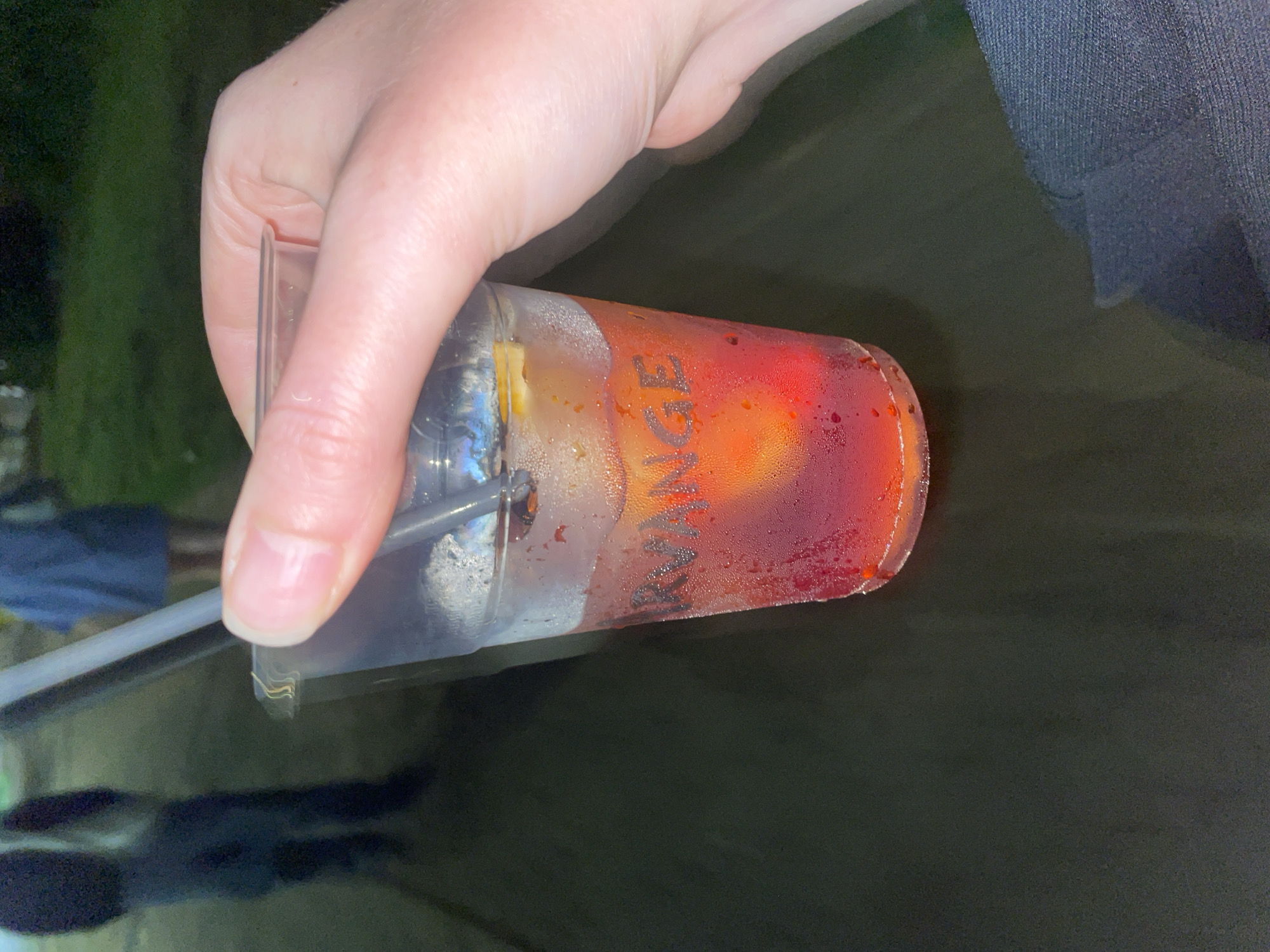 A hand holds a plastic cup containing an orange-red liquid, potentially a beverage. The cup is covered in condensation droplets and features the word "KVANGE" written on the side. A straw is inserted into the cup. The background is dark and indistinct, with blurred hints of foliage and possibly clothing. The location cannot be determined solely from this image.