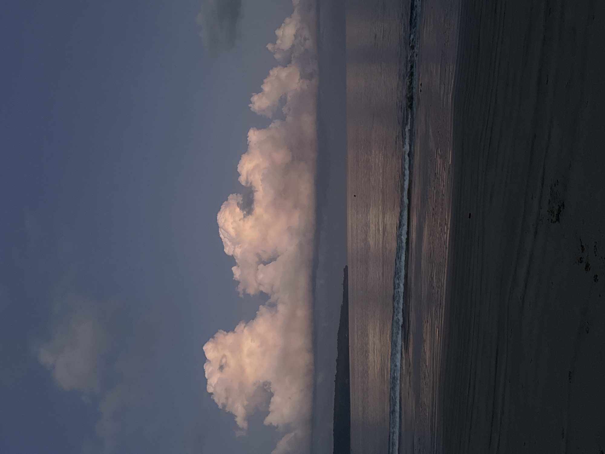 The image depicts a scene at a beach in Kuta, Indonesia, with a focus on the sky, sea, and sand. The photograph is oriented vertically.
In the sky, large cumulonimbus clouds are visible on the left side, illuminated by the setting sun, contrasting with the darker blue areas of the sky.
The sea occupies the central portion of the image. The water surface is reflective, mirroring the colors of the sky and clouds. There's a small wave breaking along the shoreline.
The sand is in the lower right of the image. The sand appears wet and dark, reflecting the light.
There are no people or animals clearly visible.