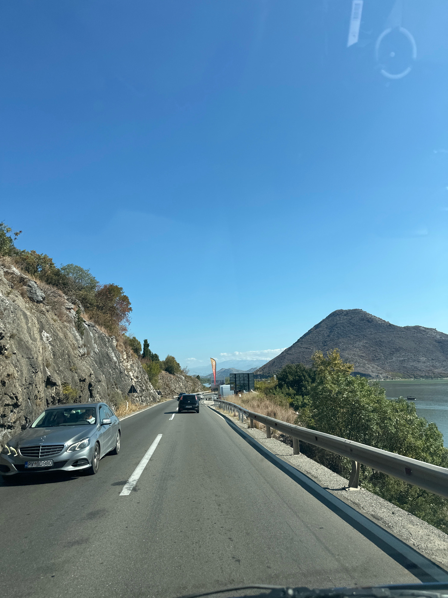 A view from inside a vehicle traveling on a paved road with a guardrail, approaching a sharp curve. To the left, a rocky, steep hillside with sparse vegetation rises. To the right, a body of water, possibly a lake, is visible, with a large, dark, conical mountain in the background. The sky is clear and blue. Several vehicles are on the road, including a silver Mercedes-Benz sedan in the foreground. The car's license plate is partially obscured but appears to read "PV BD 980". The location is the road leading to Kruševica, Montenegro.
