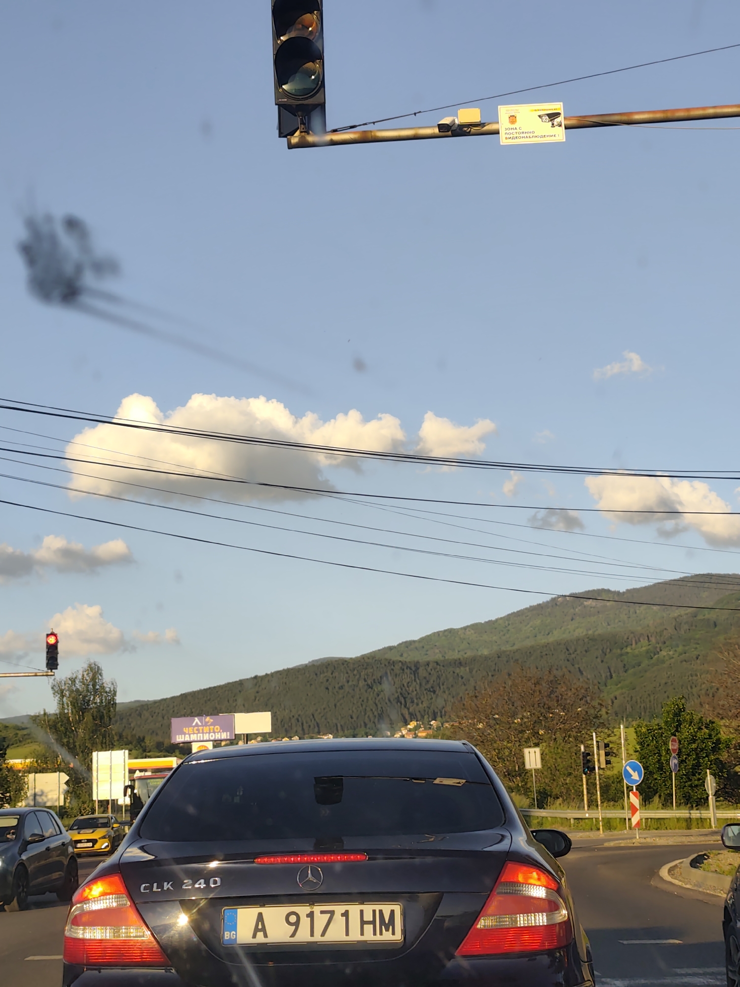The image depicts a street scene in Dragichevo, Bulgaria. The rear of a dark-colored Mercedes-Benz CLK 240 with license plate A 9171 HM (BG) is prominently displayed. The vehicle is stopped at a traffic light. Black smoke emanates from an unseen source, possibly another vehicle, in the upper-left quadrant of the image. Behind the Mercedes-Benz, a billboard with Cyrillic text is visible, along with other vehicles and a gas station. A hill or mountain covered in trees forms the backdrop. Overhead wires run across the sky. Traffic lights and road signs are visible at an intersection to the right of the Mercedes-Benz. A surveillance camera and signage are mounted on a pole above the traffic light. The sky is partially cloudy.