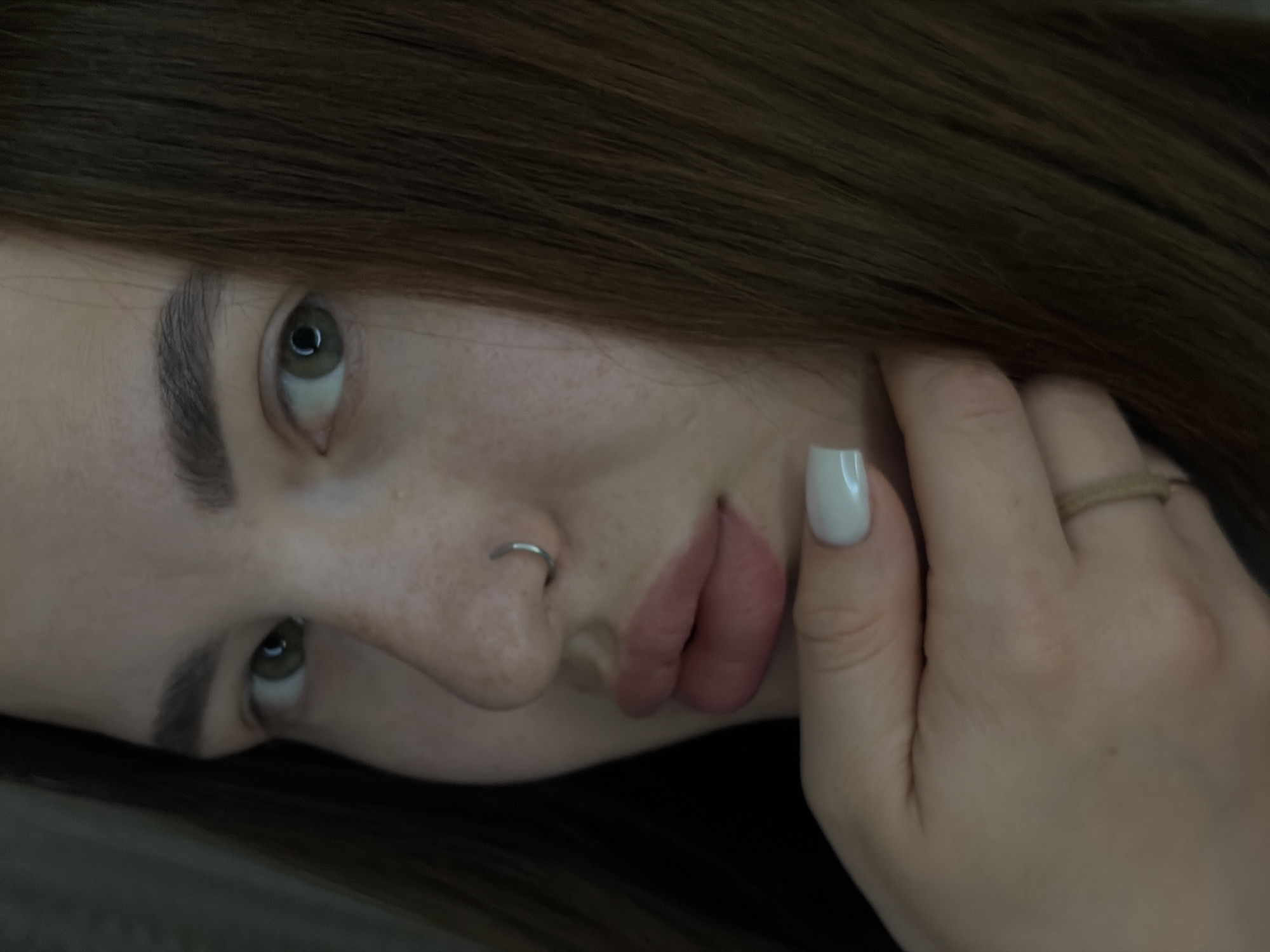 A close-up, high-angle shot of a person's face. The subject has light skin, green eyes, and brown hair that is partially visible at the top and sides of the frame. A silver ring pierces the left nostril. The person's right hand is partially visible, with a white nail and a brown ring on a finger. The background is indistinct. The location is Unknown, Unknown.