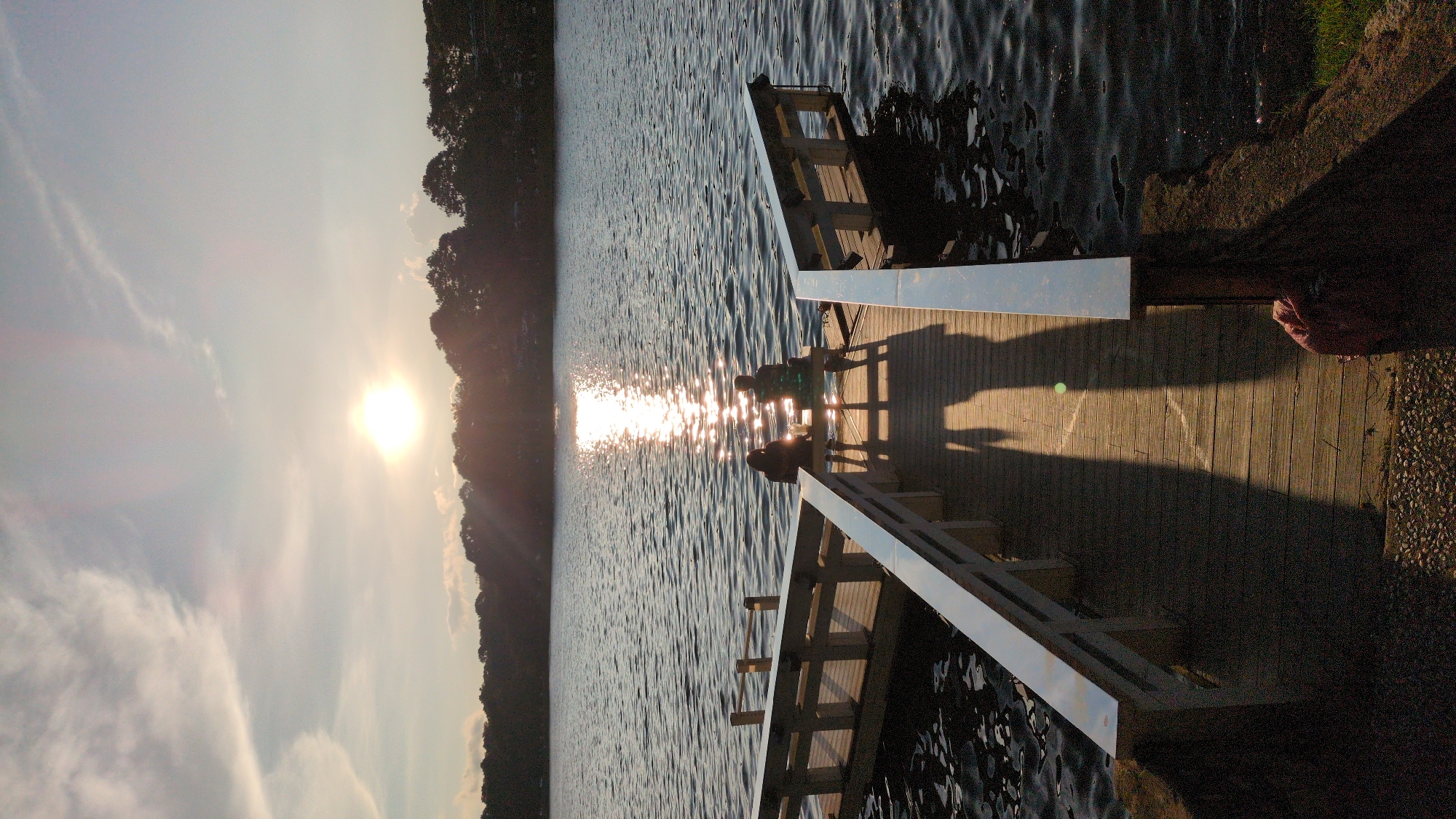 A vertical, medium-shot photograph captures a serene sunset over a lake, with two individuals seated on a wooden pier. The sun, a bright orb low in the sky, casts a shimmering path across the rippling water. A line of trees silhouetted against the horizon forms the backdrop. The pier, constructed of weathered wood, extends into the lake, with a uniquely shaped railing system. The two figures, facing away from the camera, are seated side-by-side on a bench, their shadows stretching long across the wooden planks. The water is dark and reflective, dotted with small glints of light. The atmosphere is tranquil and still. There is no information available to determine the specific location within Da Lat, Vietnam.