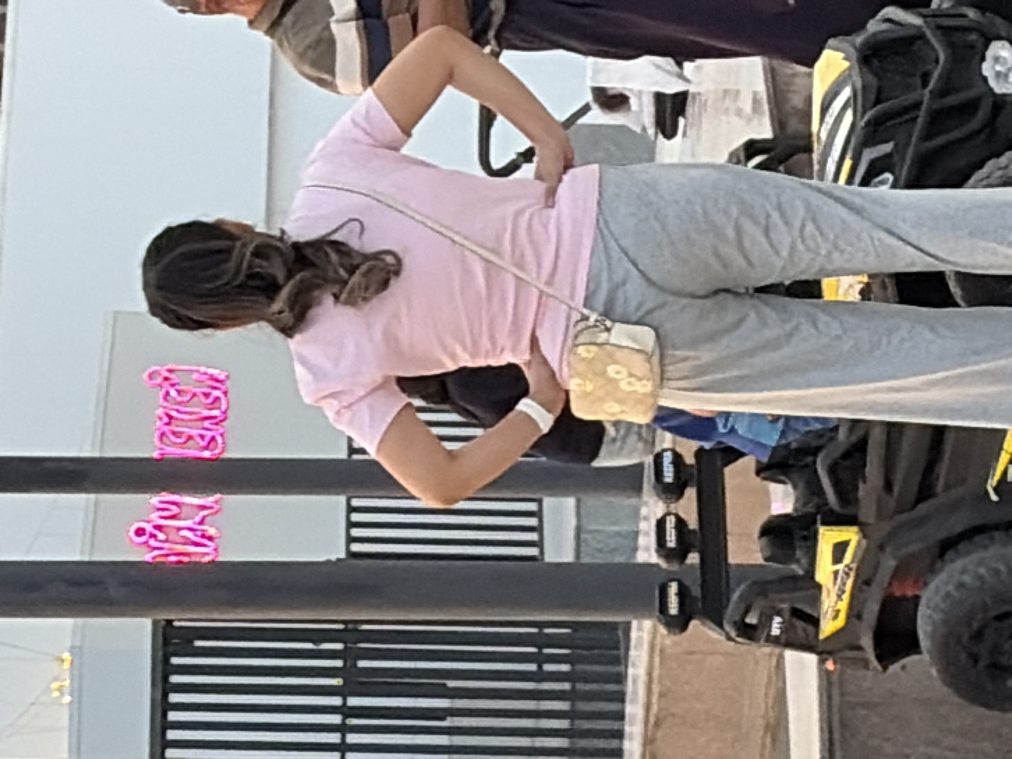 In an outdoor urban setting, a woman with dark hair pulled into a ponytail stands with her back to the camera. She is wearing a pink t-shirt and light gray sweatpants, with a small, light-colored purse with a floral pattern hanging across her body. A white wristband is visible on her left wrist. She has her hands on her hips.
In the foreground, a black and yellow toy UTV is partially visible. Three black lights with the word "KEEPER" are mounted on the roll bar of the UTV. Behind the woman, two dark gray poles are visible. In the background, a black and white striped fence is present, and beyond that, a white building with bright pink neon lettering. Another person, partially obscured, is visible to the right of the woman, wearing a striped shirt and dark pants. The lighting suggests it is daytime.
