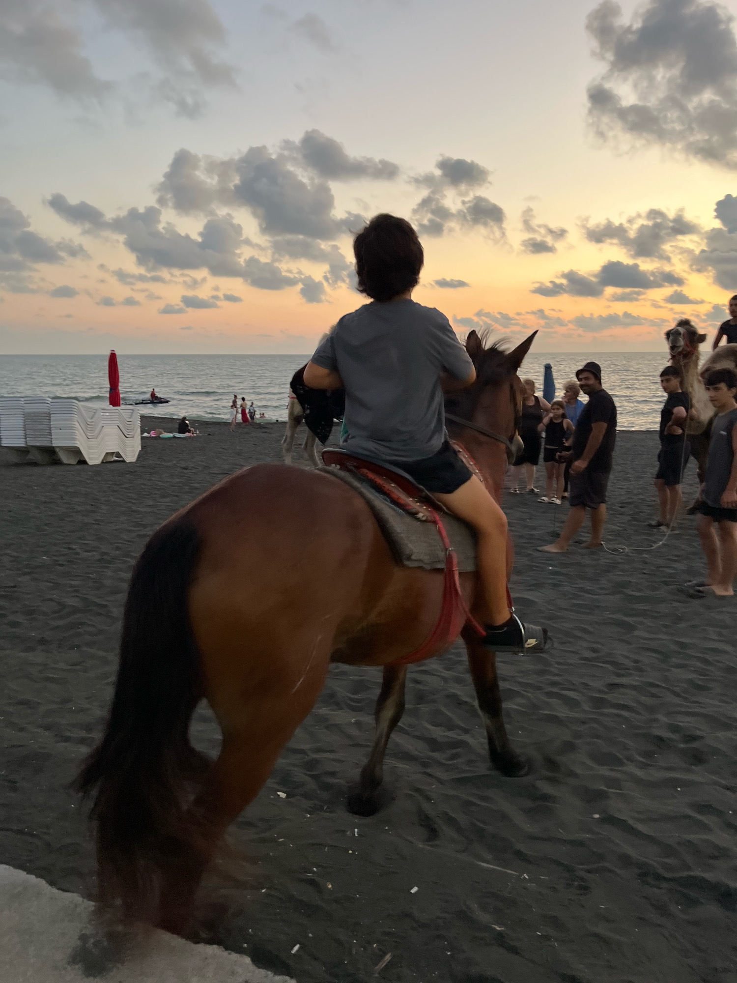 A young person rides a brown horse on a black sand beach in Ureki, Georgia. The horse is facing away from the camera, and the rider is wearing a gray t-shirt and black shorts. In the background, there are several people standing and walking, as well as two camels. The ocean is visible in the distance, with a sunset sky overhead. The scene appears to be a tourist attraction, likely offering camel and horse rides.