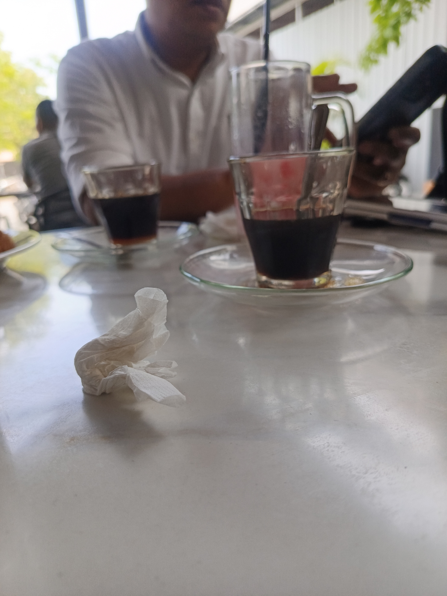 The image captures a casual daytime scene at a cafe, likely in Keuniree, Indonesia. The foreground features a light-colored, reflective table, possibly marble or laminate, with a crumpled white napkin prominently placed on its left side. On the table, two small clear glass cups filled with a dark liquid, presumably coffee, rest on clear glass saucers. One cup is closer to the viewer and in partial focus, while the other is further back and more blurred.
Behind these items, a person wearing a white collared shirt is seated, with their upper body and arm visible but out of focus. A larger clear mug, possibly containing a reddish drink or water with a straw, is positioned near this person. To their right, a hand is holding a dark smartphone, also blurred. In the background, another person is faintly visible and completely out of focus, along with vibrant green foliage, suggesting an open-air or well-lit indoor setting. The bright, natural illumination indicates it is daytime. The activity appears to be people relaxing and enjoying beverages. No visible text is present in the image.