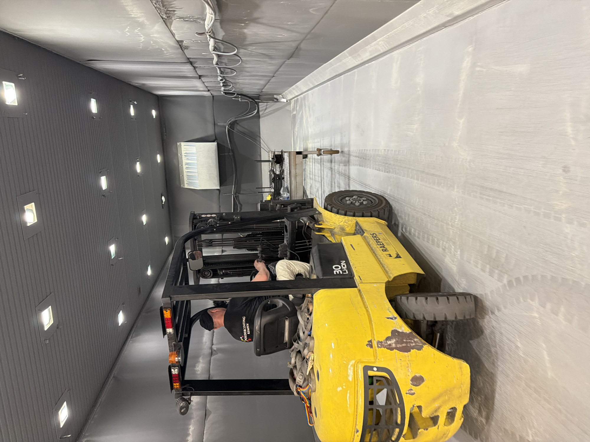 The media file depicts a man operating a yellow forklift inside a large, industrial indoor space. The forklift has "RAEPERS" and "30 LHON" visible on its side. The man is wearing a black t-shirt with text that appears to be "POEDERCONTING KEMPEN" and a black head covering. He is leaning forward, seemingly engaged in a task with the forklift's equipment.
The setting is an industrial interior, possibly a workshop or warehouse, characterized by grey, corrugated walls and a similarly colored ceiling. Numerous square lights are embedded in the wall, illuminating the space. A large, white ventilation unit is visible on the left wall. The floor is concrete and shows tire marks from vehicles. The overall environment appears to be functional and industrial. The lighting suggests an indoor setting, and there are no indicators of the time of day or weather conditions. The scene is focused on the operation of the forklift and the man's involvement with it.