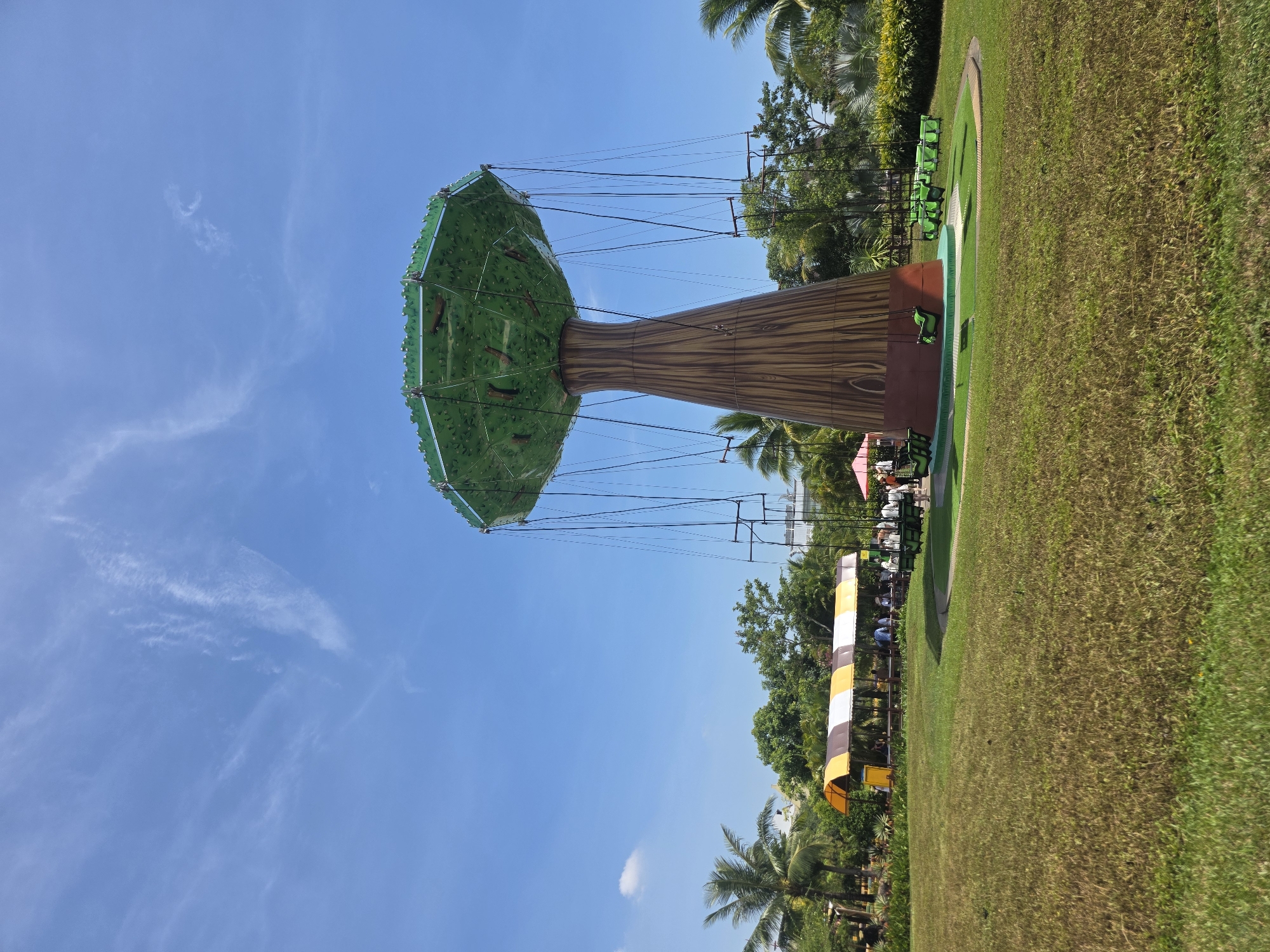The image captures a whimsical amusement park attraction, likely a swing ride, designed as a stylized tree. The "tree trunk" is brown with wood grain detailing, topped with a wide, green "foliage" canopy from which swing seats are suspended.
The ride is situated on a grassy field under a clear blue sky with scattered cirrus clouds. In the background, lush greenery and palm trees suggest a tropical setting. To the right of the ride, there's a fenced area with green seating. In the distance, a shaded area with a striped awning indicates a waiting area or concessions. The location is likely the Asia Park in Đà Nẵng, Vietnam, judging by the architectural style and tropical landscaping. There are indistinct figures under the awning, possibly visitors waiting for their turn on the ride. No specific actions or interactions are discernible due to the distance and image angle.