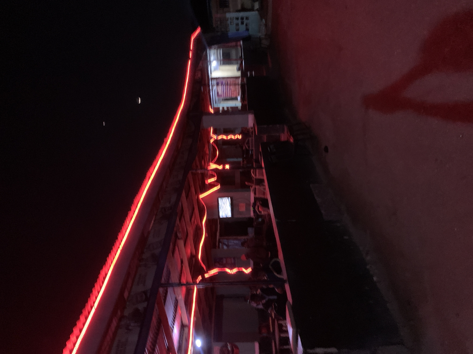 At night in New Stadium, Nigeria, a multi-story building is illuminated with red neon lights outlining its top and various sections. A crescent moon and a small light source are visible in the dark sky to the left of the building. A road stretches along the right side of the building. People are visible inside and on the balconies of the building.
