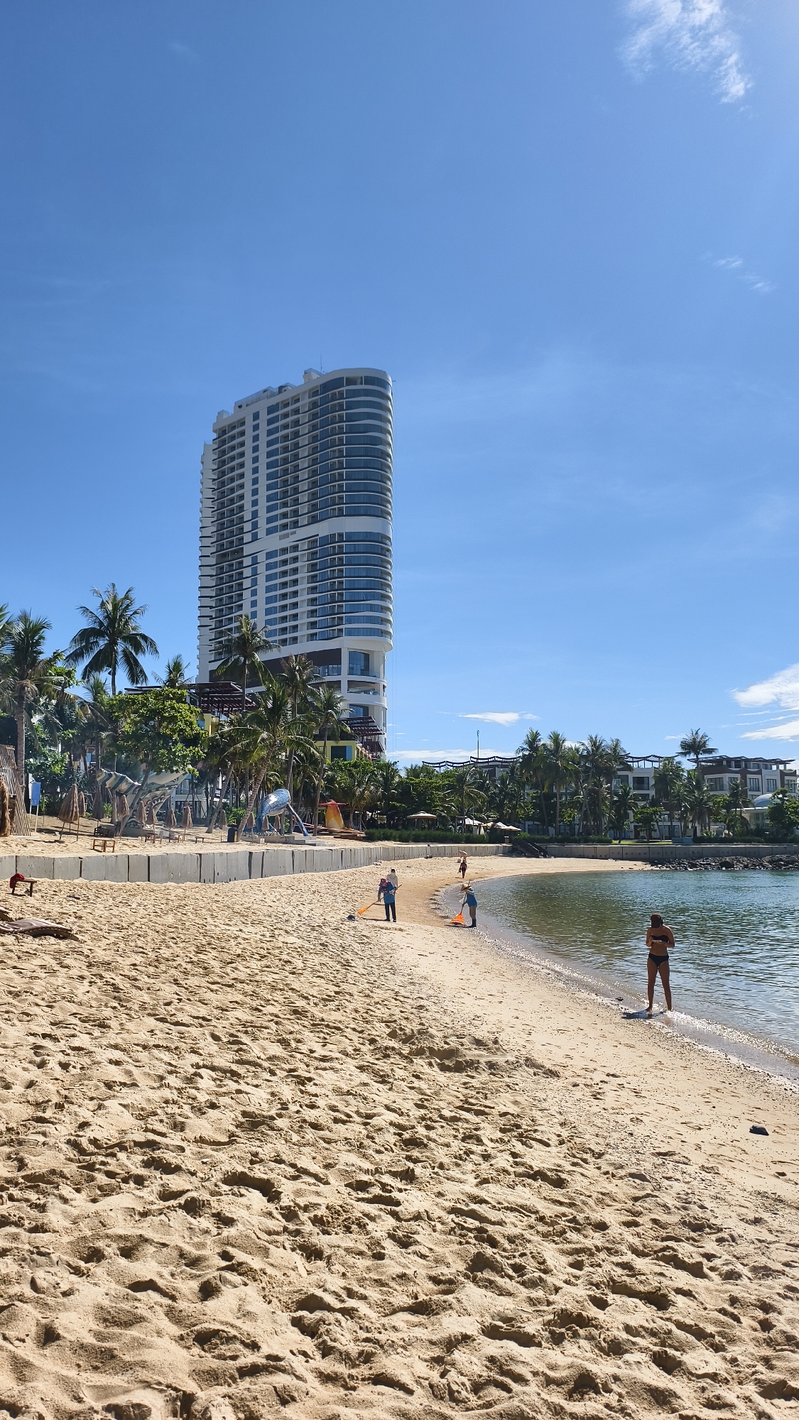 This image captures a sunny day at a beach in Nha Trang, Vietnam, with a prominent tall, curved white building in the background. The beach itself is sandy with some footprints and scattered debris. Several people are present. In the mid-ground, two individuals, one wearing a blue shirt and shorts and the other a blue top and light-colored shorts, are using orange shovels on the sand, appearing to be engaged in some activity. Further down the beach, closer to the water, a person wearing a black bikini stands at the edge of the sea. The water is calm and appears to be an ocean or large bay. Behind the beach, there is a line of palm trees, some beach umbrellas, and further structures, suggesting a resort or developed coastal area. A concrete barrier runs along the edge of the beach in some sections. The sky is clear and blue with a few wispy clouds.