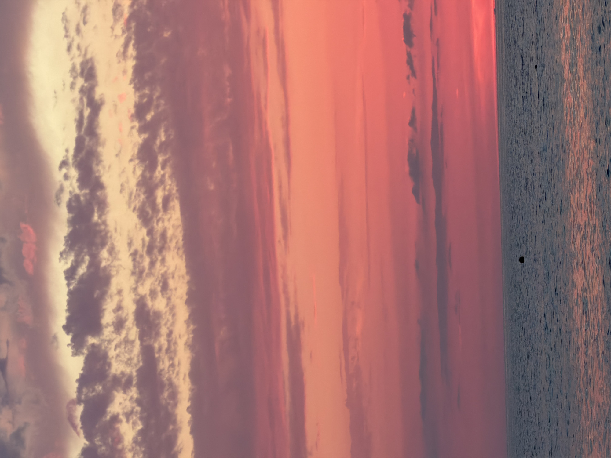A sunset over a body of water. The sky is filled with a gradient of colors, transitioning from a pale yellow and pink at the horizon to a deep magenta and purple higher up. Wispy clouds are scattered across the sky, catching the light and adding texture. The water below is a deep blue, with subtle ripples reflecting the colors of the sunset. A small, dark object, possibly a buoy, is visible in the distance on the surface of the water. The overall scene is serene and picturesque.