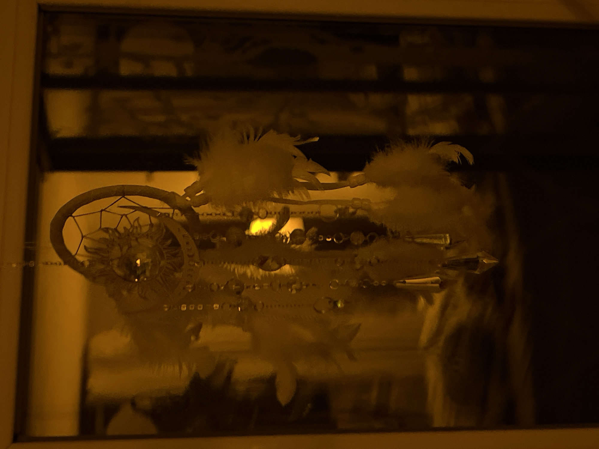 A dreamcatcher, adorned with feathers and beads, hangs in front of a window in an indoor setting. The lighting in the scene is dim and casts a warm, yellow hue over the objects. The background, visible through the window, appears to be a dark, indistinct interior space with faint reflections. The dreamcatcher is the central focus, with its intricate design and delicate embellishments visible.