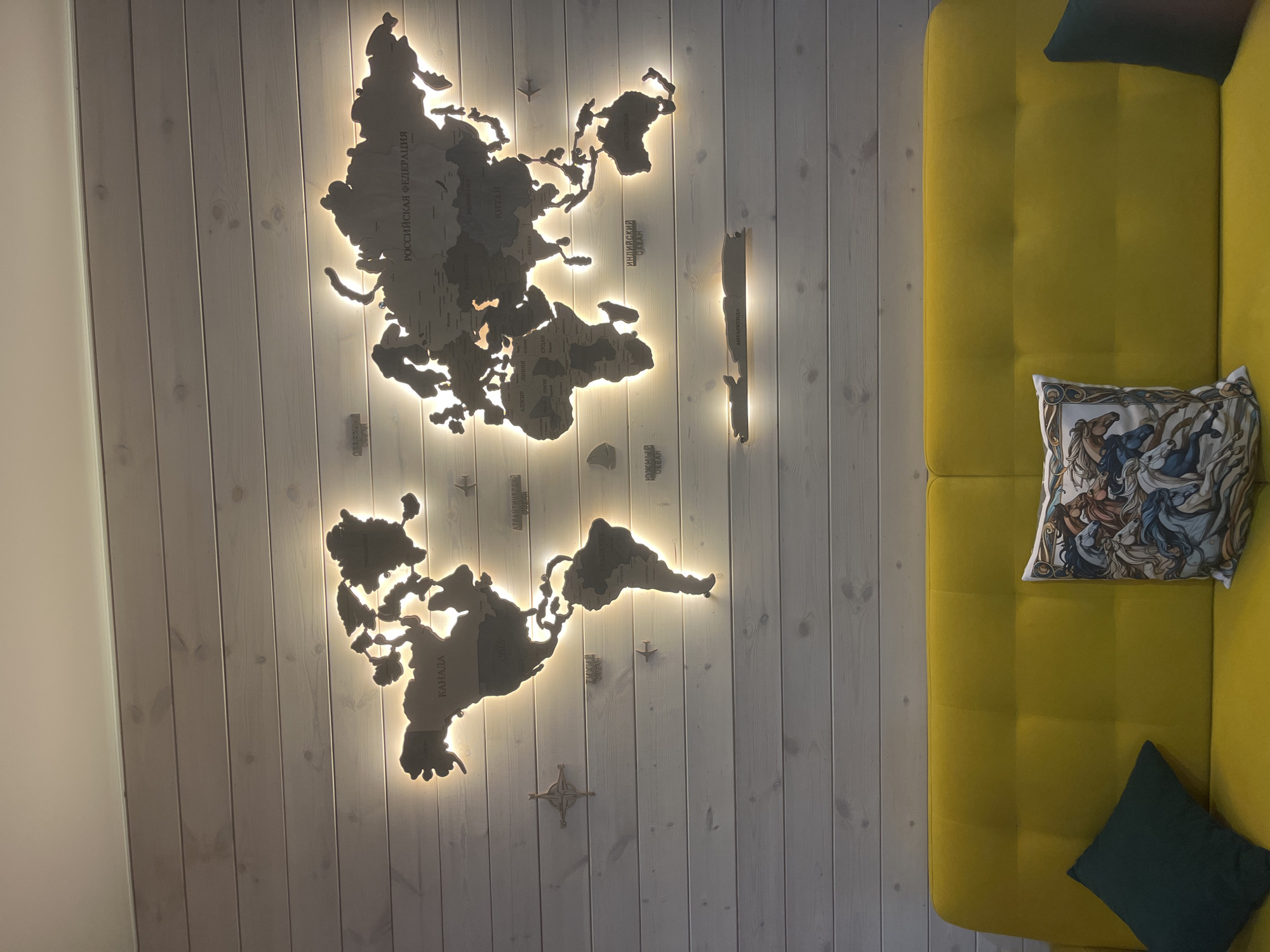 From a high-angle perspective, the scene presents a yellow sofa with two cushions (one patterned with horses, the other a solid dark green) positioned beside a wall decorated with a wooden, backlit world map. The wall has vertical white planks. The location is Новая Чернушка, Russia. There are no people visible.