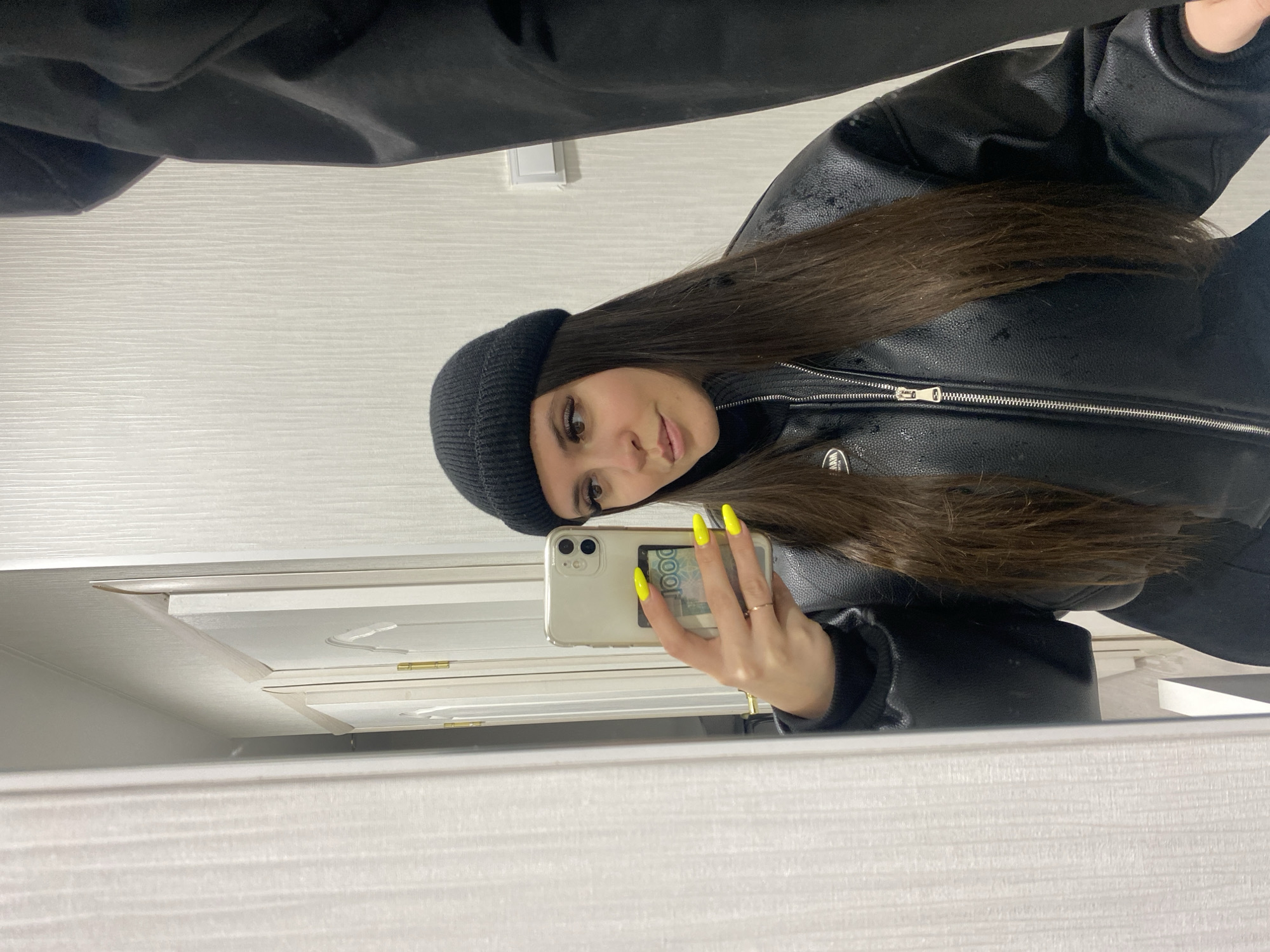 The image captures a young woman taking a mirror selfie indoors. She is positioned to her right, looking directly at her reflection in the mirror with a neutral expression. Her long, straight dark brown hair falls over her shoulders and down her back. She wears a black knitted beanie and a black leather jacket with a visible silver zipper and what appear to be small water droplets or reflective specks on its surface.
She holds a light-colored smartphone in her left hand, which is encased in a clear cover. A 1000 Russian Ruble banknote, with "1000" and partial text "РОО" visible, is tucked into the phone case. Her fingernails are painted a vibrant neon yellow. A simple golden ring is worn on her left ring finger.
The background reflected in the mirror suggests an interior setting, likely a room in Chaikovsky, Russia, as indicated by the currency and context. Light-colored walls or furniture with horizontal textures are visible, along with parts of what appears to be a white door or cabinet featuring decorative moldings and golden hinges. Above the reflected scene, a dark, draped fabric or clothing item is visible. The lighting is bright and even, indicating an indoor environment.