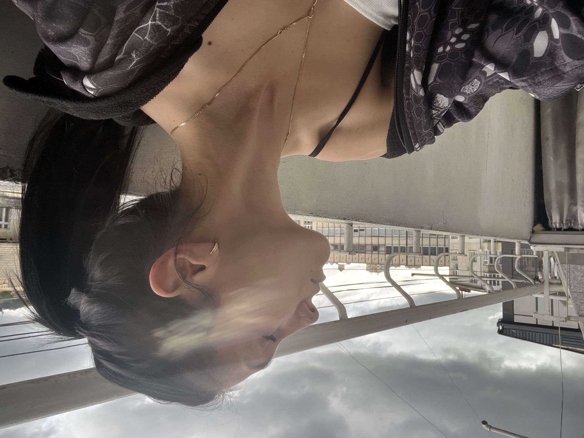 An image, rotated on its side, displays a close-up profile view of a person, likely female, from the chest up. The person has dark hair pulled back and is looking to the left of the frame (when upright), with eyes slightly downcast. She wears a dark, patterned zip-up jacket over a white top, with a thin gold chain necklace draped around her neck. A small, shiny hoop earring is visible in her right ear. A faint, soft haze or reflection partially obscures her face.
The background suggests an outdoor urban setting, possibly a balcony or rooftop, as indicated by a prominent white railing that runs horizontally across the lower part of the frame. Beyond the railing, several multi-story buildings with numerous windows are visible. The sky is overcast with thick grey clouds, suggesting a cloudy day or approaching evening. There is no discernible activity other than the person's contemplative posture. No text is visible in the image.