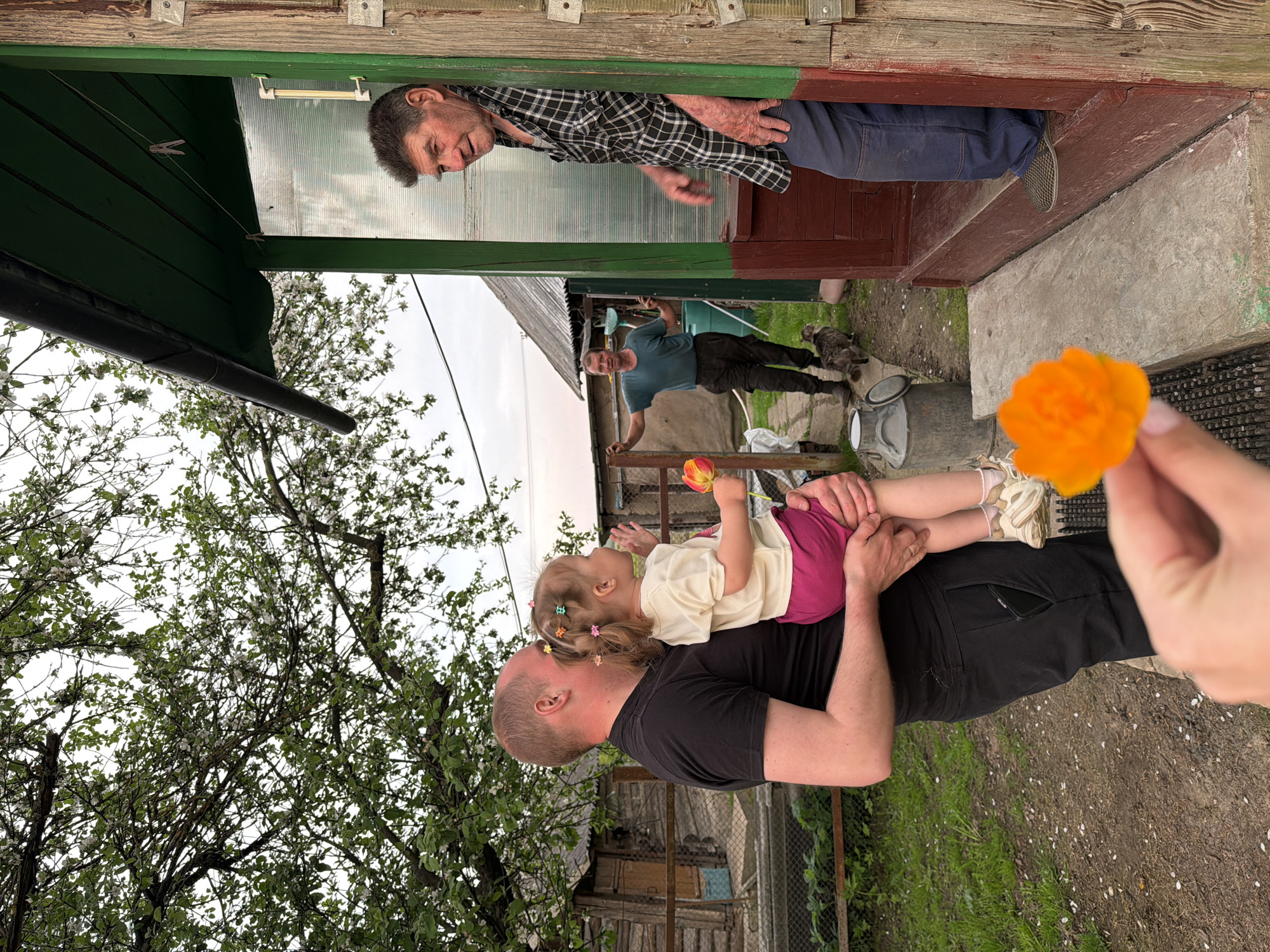 A photo taken in Спас-Заулок, Russia, shows a man holding a young child who is reaching for an orange flower held in someone's hand. In the background, another man is standing next to a man looking out a window. A cat is also visible on the ground. The setting appears to be a rural residential area.
