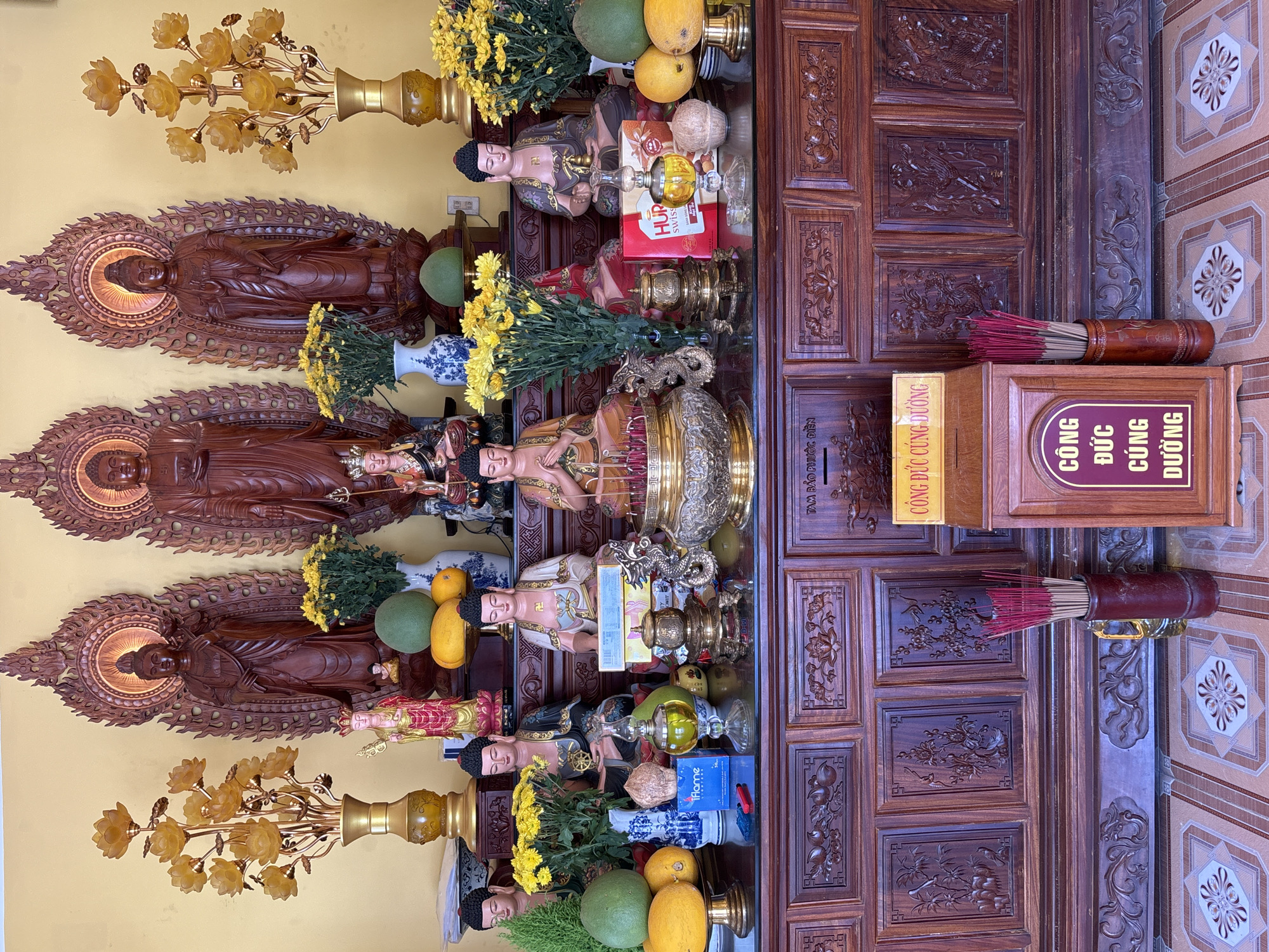 The image depicts an altar in what appears to be a Buddhist temple or a shrine, likely located in Nha Trang, Vietnam. The altar is constructed of dark, intricately carved wood, with multiple shelves displaying various religious artifacts.
Dominating the upper level are three large, wooden Buddha statues, each with a distinct posture and intricate carvings. Flanking these central figures are two tall, golden floral lamps with yellow, lotus-shaped shades. Smaller statues of deities or monks are also present on this level, some standing and others seated.
The middle shelf is laden with offerings and decorative items. These include large, green, spherical fruits (likely pomelos), yellow fruits, bouquets of yellow chrysanthemums in blue and white vases, several small Buddha statues, incense burners, and various containers. A prominent golden incense burner sits at the center, adorned with a dragon sculpture. To the right of this burner are red and white boxes, possibly containing religious offerings or incense. A glass lamp with yellow oil is also visible.
The lower level of the wooden structure features carved panels depicting natural scenes, possibly flowers and birds. In front of this structure, on the floor, stands a wooden donation box with a yellow sign that reads "CÔNG ĐỨC CÚNG DƯỜNG" (Donation). To the left of the donation box, a brown cylindrical vase holds a bundle of red incense sticks.
The overall scene is one of reverence and devotion, characterized by the abundance of religious icons, offerings, and the ornate wooden furnishings. The backdrop is a plain yellow wall. The floor is tiled with a geometric pattern of light and darker colors.