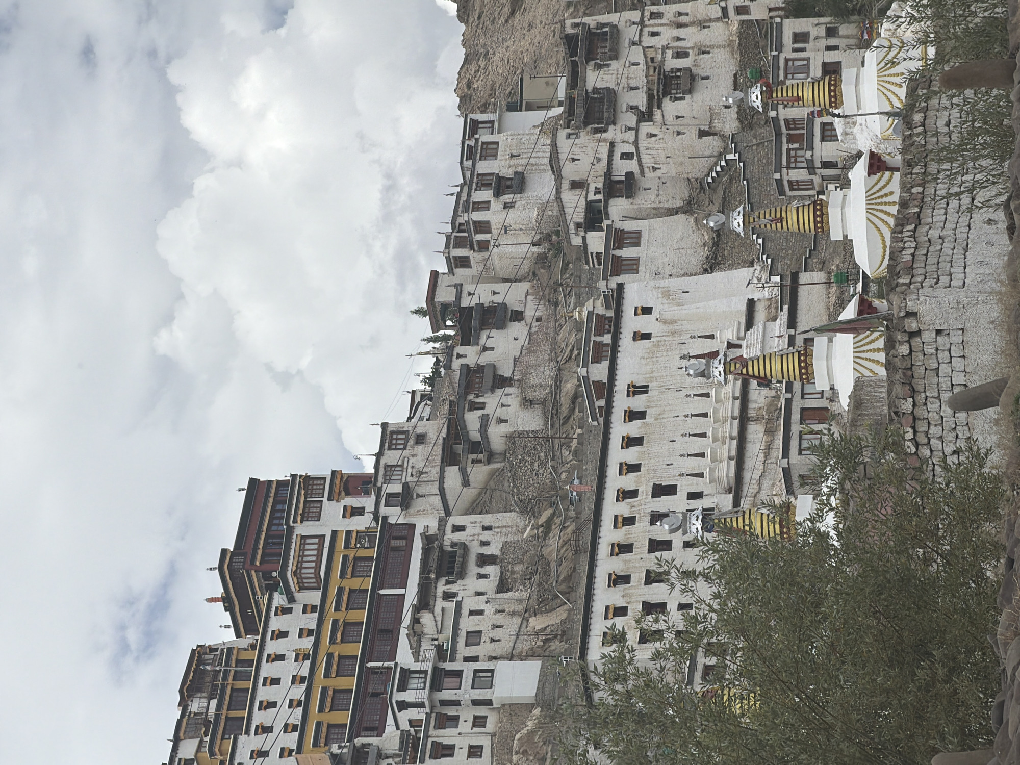 The image displays a view of the Thiksey Monastery, a prominent Buddhist monastery in Ladakh, India. The monastery is built on a hilltop, with its white-washed buildings cascading down the slope. The architecture is characterized by multi-storied structures with flat roofs and numerous windows. In the foreground, several stupas, traditional Buddhist burial mounds, are visible. They are white with gold embellishments and are topped with spire-like structures. Green foliage from trees partially obscures the lower part of the monastery. The sky above is cloudy, with patches of blue peeking through. The overall scene depicts a serene and majestic religious complex set against a natural landscape.