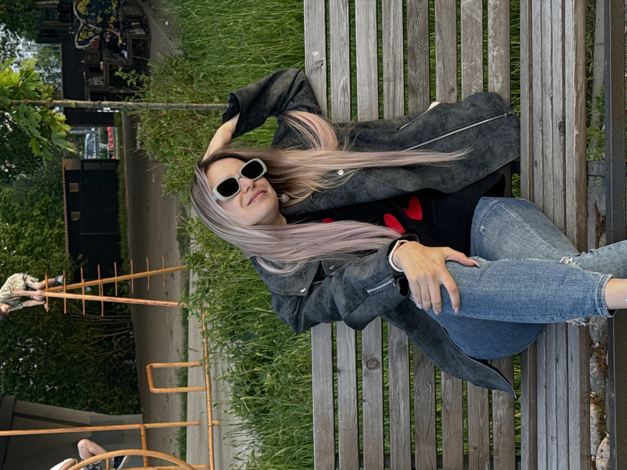 A young woman with long, light-toned hair (possibly lavender or pinkish) and stylish rectangular sunglasses is lying on her back on a rustic wooden park bench. She is wearing a dark grey or black textured jacket over a black top adorned with red heart patterns, and light blue ripped jeans. Her left arm is bent, with her hand resting behind her head, and her right hand is casually placed on her knee. She appears relaxed, possibly smiling faintly behind her sunglasses.
The bench is composed of weathered wooden slats. Behind her and to the side, there is lush green grass and various trees and bushes, suggesting a park or green recreational area. In the mid-ground of the background, a paved path is visible, running alongside more greenery and what appears to be a small dark building or structure. Further back, obscured by trees, some urban elements like buildings and possibly a vehicle can be vaguely seen.
To the left in the background, a person in camouflage-patterned clothing is interacting with a tall, wooden and metal playground or climbing structure. Another person's legs and feet are partially visible at the bottom left, also near a similar metal structure.
The scene takes place during the daytime, with soft, diffused lighting, suggesting an overcast day or late afternoon. The overall atmosphere is tranquil and casual. No discernible text is visible in the image. The setting appears to be a public park or recreational ground in an urban or suburban environment, consistent with locations like Saint Petersburg, Russia.