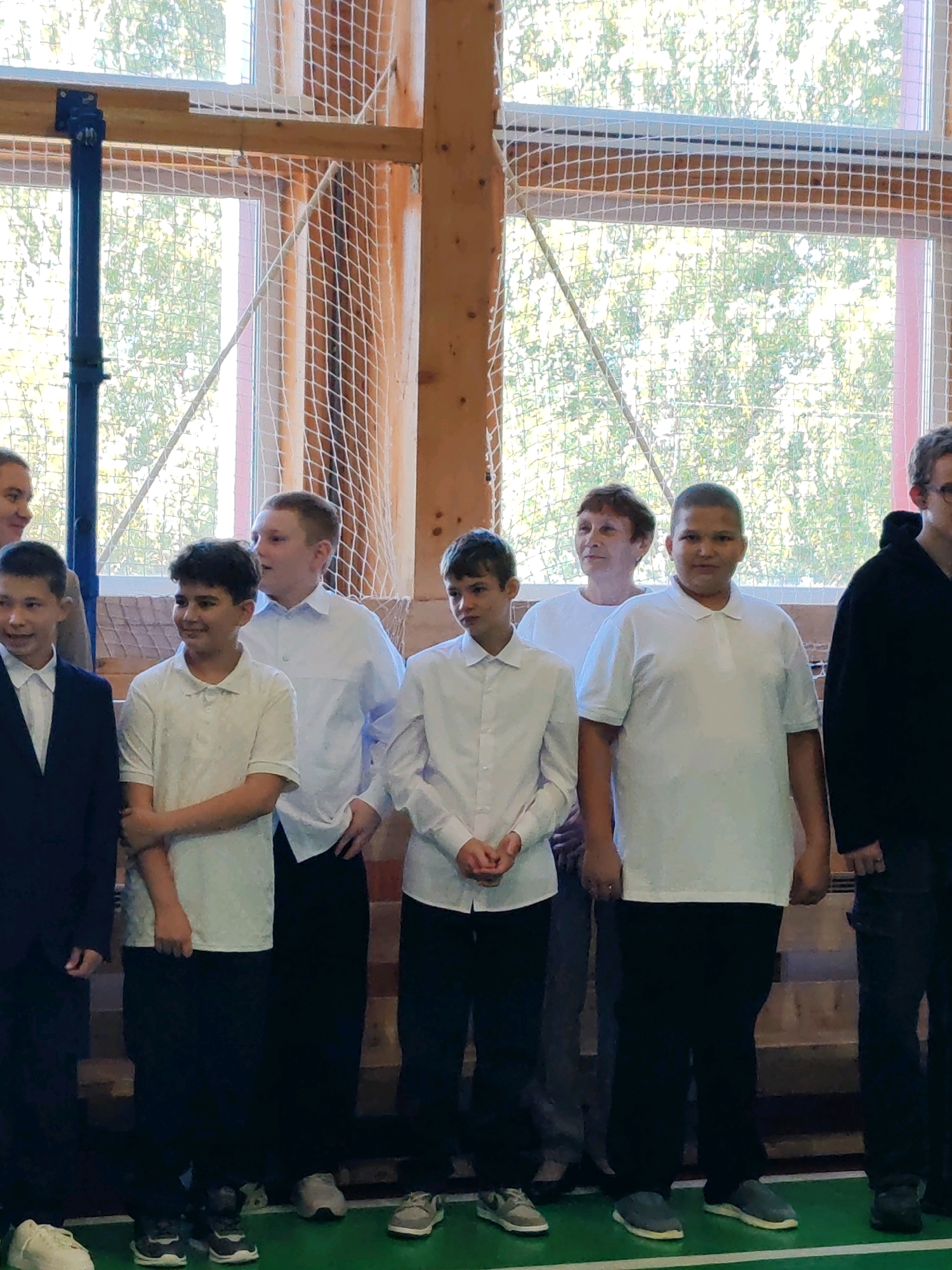 A group of people, mostly young males, stand in a gymnasium, likely at a school. The room features wooden walls, large windows with netting, and a blue pole, possibly part of a sports apparatus. The individuals are casually dressed, with some wearing white shirts and dark pants, while others are in suits or hoodies. The overall atmosphere suggests an indoor event or gathering, possibly a school sports day or assembly. The individuals are standing in a line facing forward, with some looking towards the camera and others looking away. The lighting is natural, coming from the windows, and the scene is captured from a medium distance, showing the upper bodies and lower bodies of the people. The floor is green, indicating a sports surface. This scene is located in the city of Chukhlem, Russia, though the specific building or institution is not identified.