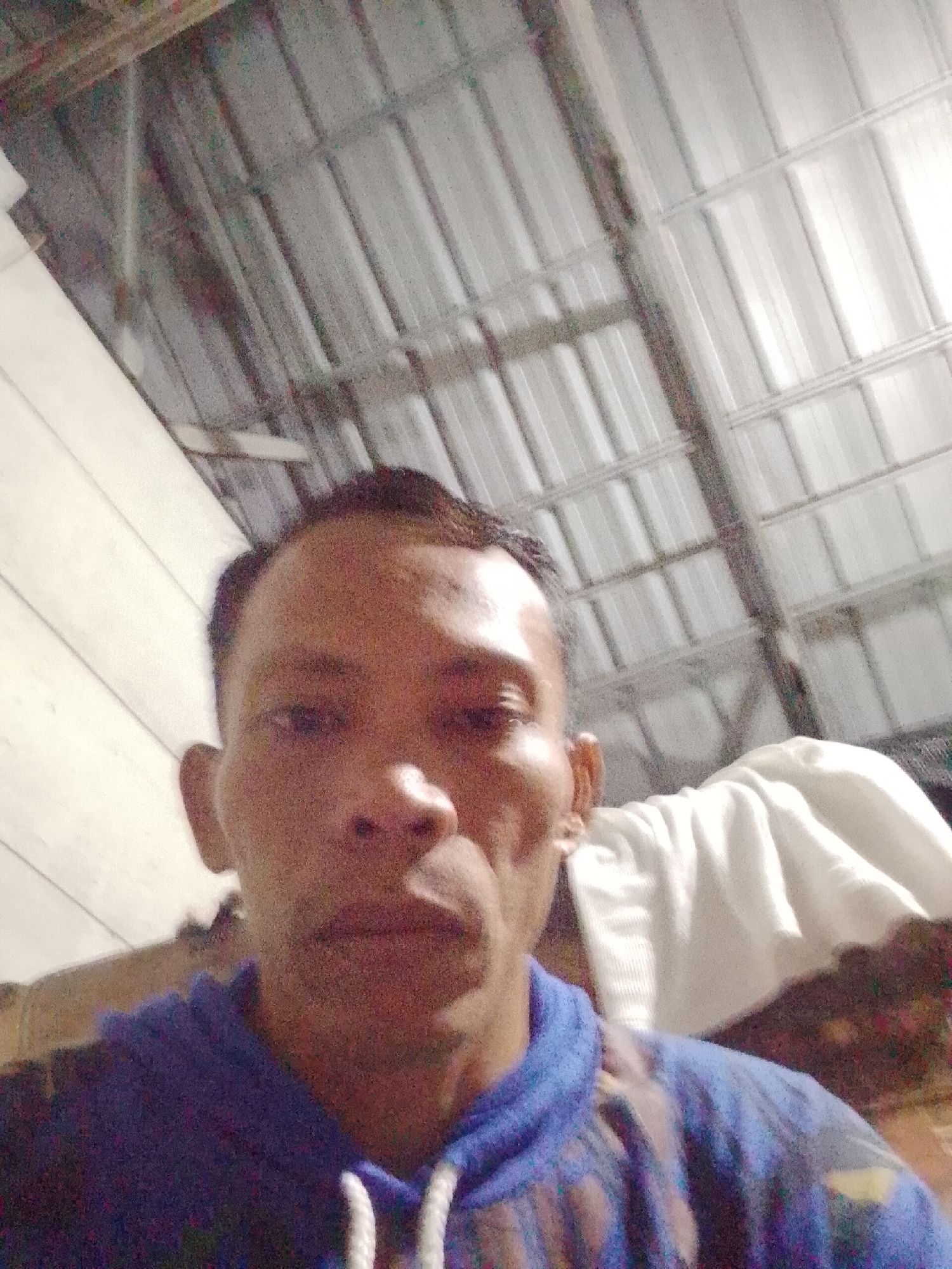 The image shows a man in a blue and patterned hoodie, likely in an indoor location, evidenced by the corrugated metal sheet ceiling with supporting framework. A white cloth or garment is visible in the background. The photo was taken in Bukit Ketok, Indonesia.