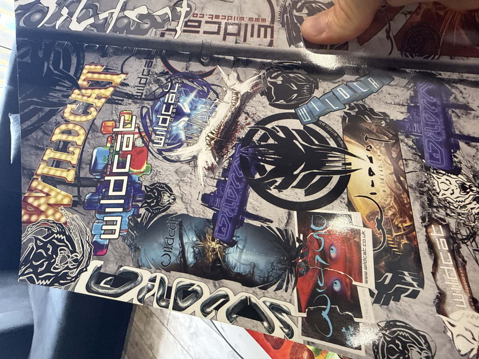 The image displays a surface covered in stickers and labels, most of which prominently feature the word "Wildcat" in various stylized fonts and designs. Some stickers include the URL "www.wildcat.co.uk," and there are also graphic images of cat heads or abstract representations of cats. Other prominent elements include what appear to be futuristic weapons or structures rendered in different colors and styles. There is a hand visible in the upper right corner of the image, partially obscuring the surface. Based solely on the image, there is no information indicating the location of Sofia, Bulgaria.