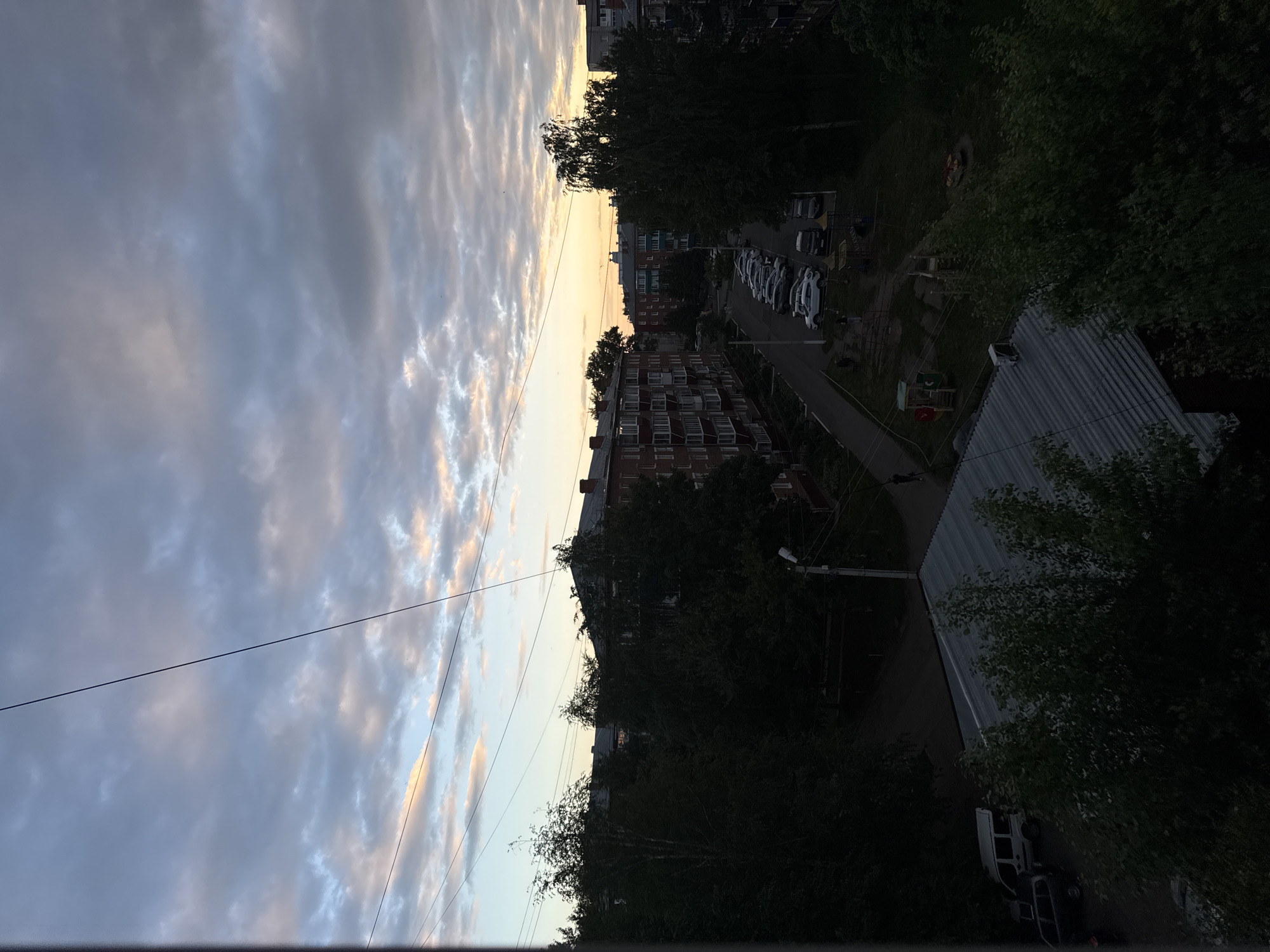 The image depicts an evening scene in the city of Sarapul, Russia, likely from a high vantage point such as a balcony or window. The sky is dominated by dramatic clouds, with a band of soft, warm light on the horizon suggesting either sunrise or sunset. Several thin wires crisscross the frame, obscuring parts of the sky.
Below the sky, apartment buildings with brick facades are visible, some with balconies and windows. Dense trees, mostly dark green, are interspersed between the buildings and in the foreground, casting shadows. A street or road runs through the scene, with parked cars visible, including what appears to be a white van. A lamppost stands near the road. Further into the scene, a playground with slides and a merry-go-round can be seen. A few indistinct figures, likely pedestrians, are present on the ground. The overall lighting suggests the twilight hours, with a transition from the fading daylight to approaching darkness.