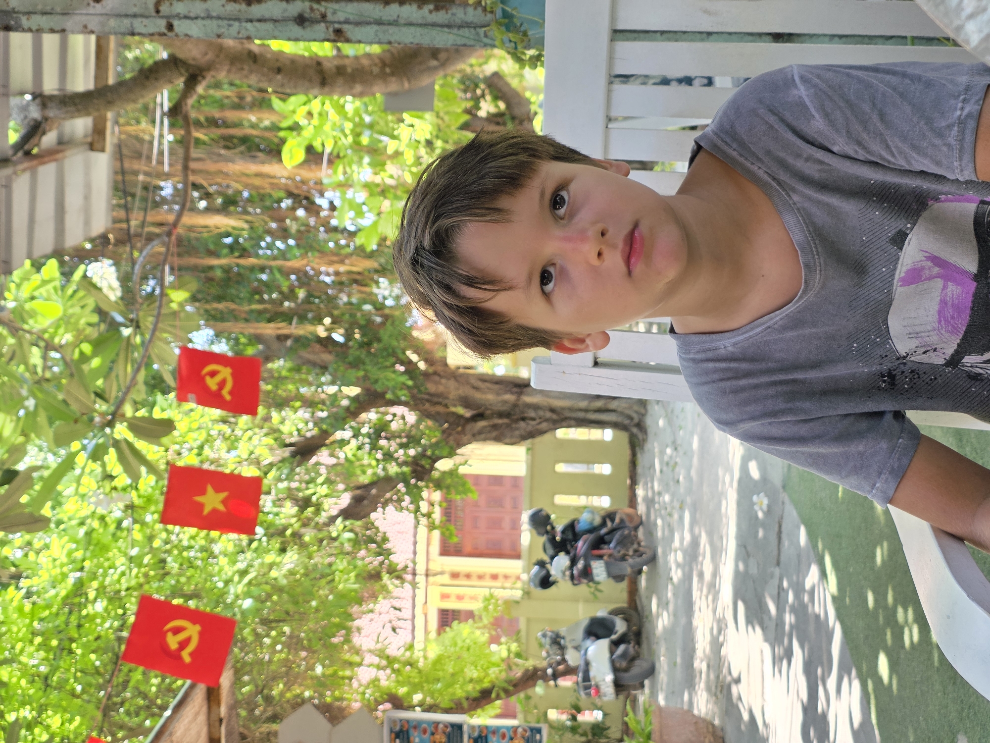 A young boy with brown hair and brown eyes sits on a white wooden bench, looking upwards. He is wearing a grey t-shirt. In the background, there are trees with lush green foliage. Red flags with yellow symbols hang from a wire strung between trees. To the left of the boy, two motorbikes are parked on a paved area with some green artificial turf. Behind the motorbikes is a building with yellow walls and red doors. The scene appears to be an outdoor seating area of a restaurant or cafe in Mui Ne, Vietnam, given the presence of the motorbikes and the general tropical atmosphere.