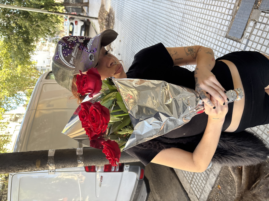 A young woman with tattoos holds a bouquet of red roses wrapped in silver foil. She wears a black crop top and shorts, plus a baseball cap. She stands on a sidewalk beside a white van. Her gaze is directed toward the camera, and the overall scene appears to be outdoors during the day.