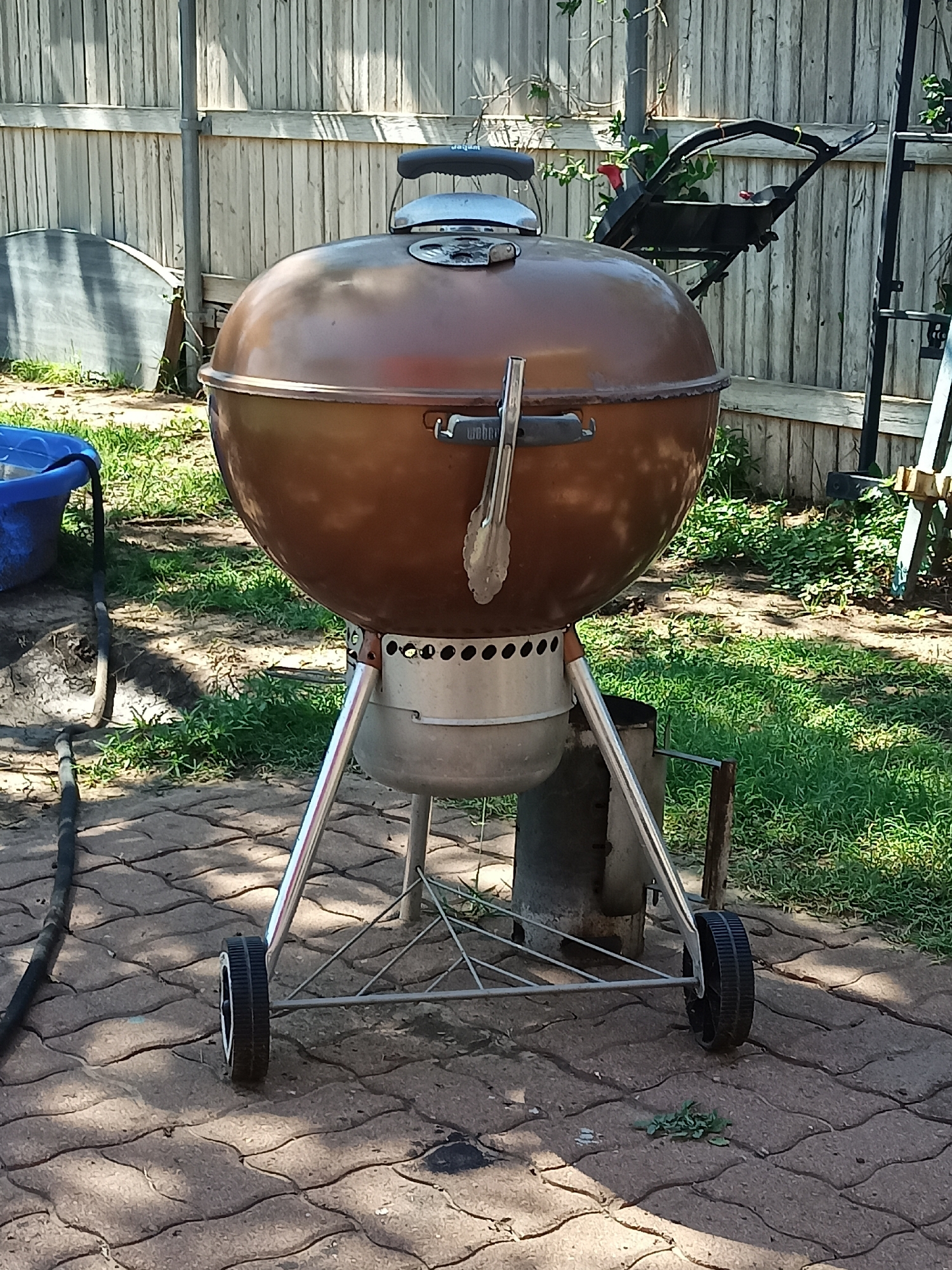 A copper-colored Weber kettle grill stands on a brick patio in a backyard. The grill is the central focus of the image and has a large dome-shaped lid, a silver handle on top, and a silver grilling tool clipped to the side. The grill sits on a silver frame with two wheels visible at the bottom. Behind the grill is a wooden fence and some greenery. To the left of the grill, a blue plastic container is partially visible, and a black hose lies on the ground. The scene appears to be in a residential area, likely in Amarillo, United States of America, given the commonality of such backyards and grills in that region. The lighting suggests it is daytime, with sunlight casting shadows on the patio.