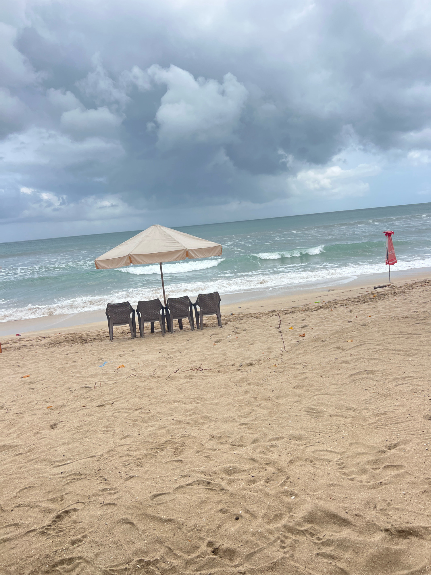 The image depicts a beach scene in Kuta, Indonesia, under a cloudy sky. In the foreground, textured sand shows footprints and scattered small debris. A row of four dark plastic beach chairs sits on the sand, facing the ocean. Behind the chairs, a light-colored umbrella is open, providing shade. The ocean in the background features moderate waves and a greenish-blue hue. To the right of the umbrella, a red umbrella or flag on a pole stands closer to the water's edge. The overall atmosphere suggests an overcast day at the beach.