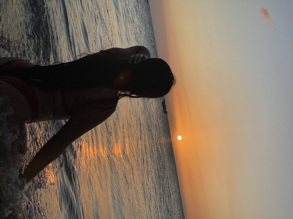 A silhouetted woman in a bikini stands at the shoreline as the sun sets. Her dark, long hair extends down her back. Light from the setting sun reflects on the water. A small boat is visible further out on the water, near the horizon. The sky transitions from orange near the sun to lighter hues above.