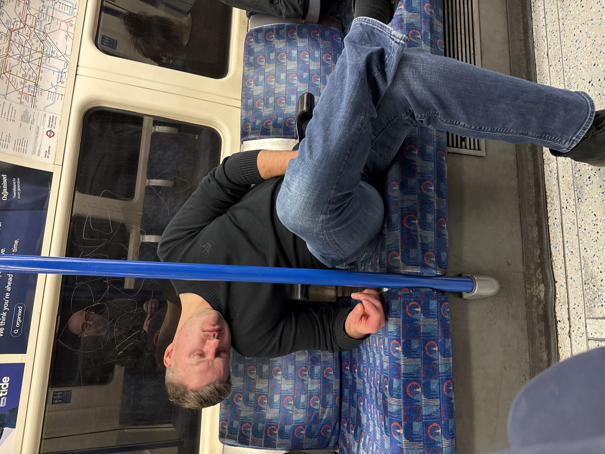 The image shows a man sleeping across multiple seats on a London Underground train. He is wearing a black sweater and blue jeans, and has his legs stretched out, resting his feet on the seat next to the aisle. The train car has the typical blue and red patterned seating, and the walls have route maps and advertisements. Another person is partially visible in the reflection of the train window.