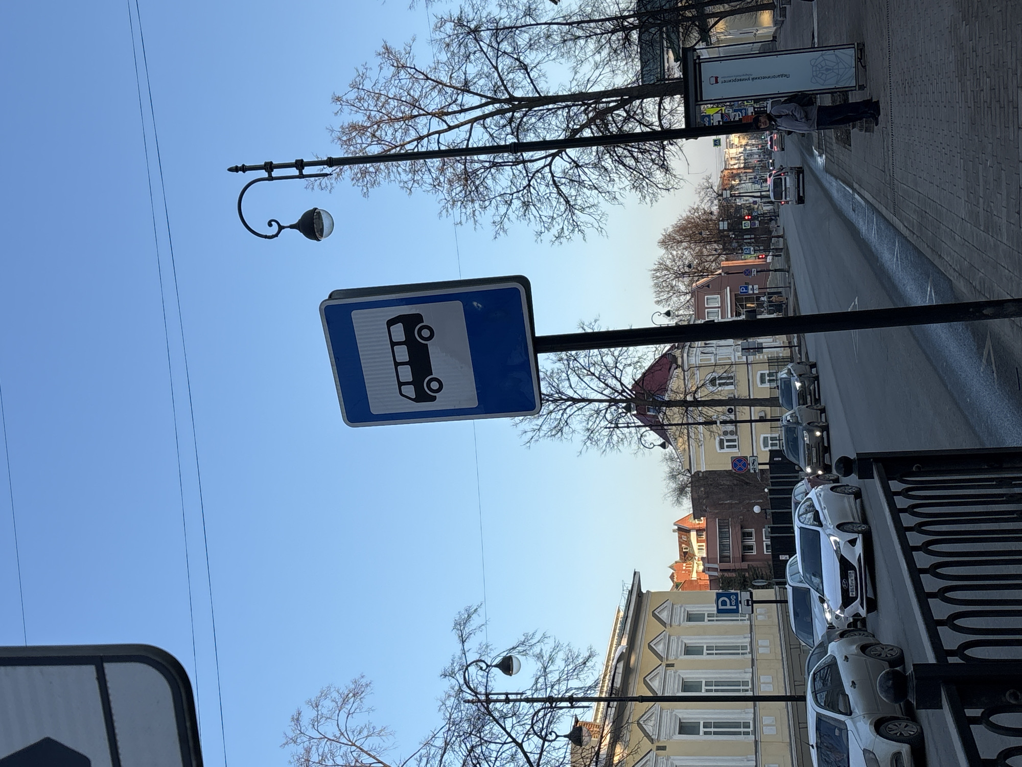 The image is taken in Perm, Russia, at a bus stop with a distinctive blue and white bus sign. The bus stop is located on a street lined with buildings of various heights and colors, mostly in pastel shades. A row of parked cars is visible along the curb. A person stands near a sign on the sidewalk, partially obscured. Street lamps with curved arms and globe-shaped lights are positioned along the street. Bare trees are interspersed among the street furniture and buildings. The sky is a clear, pale blue.