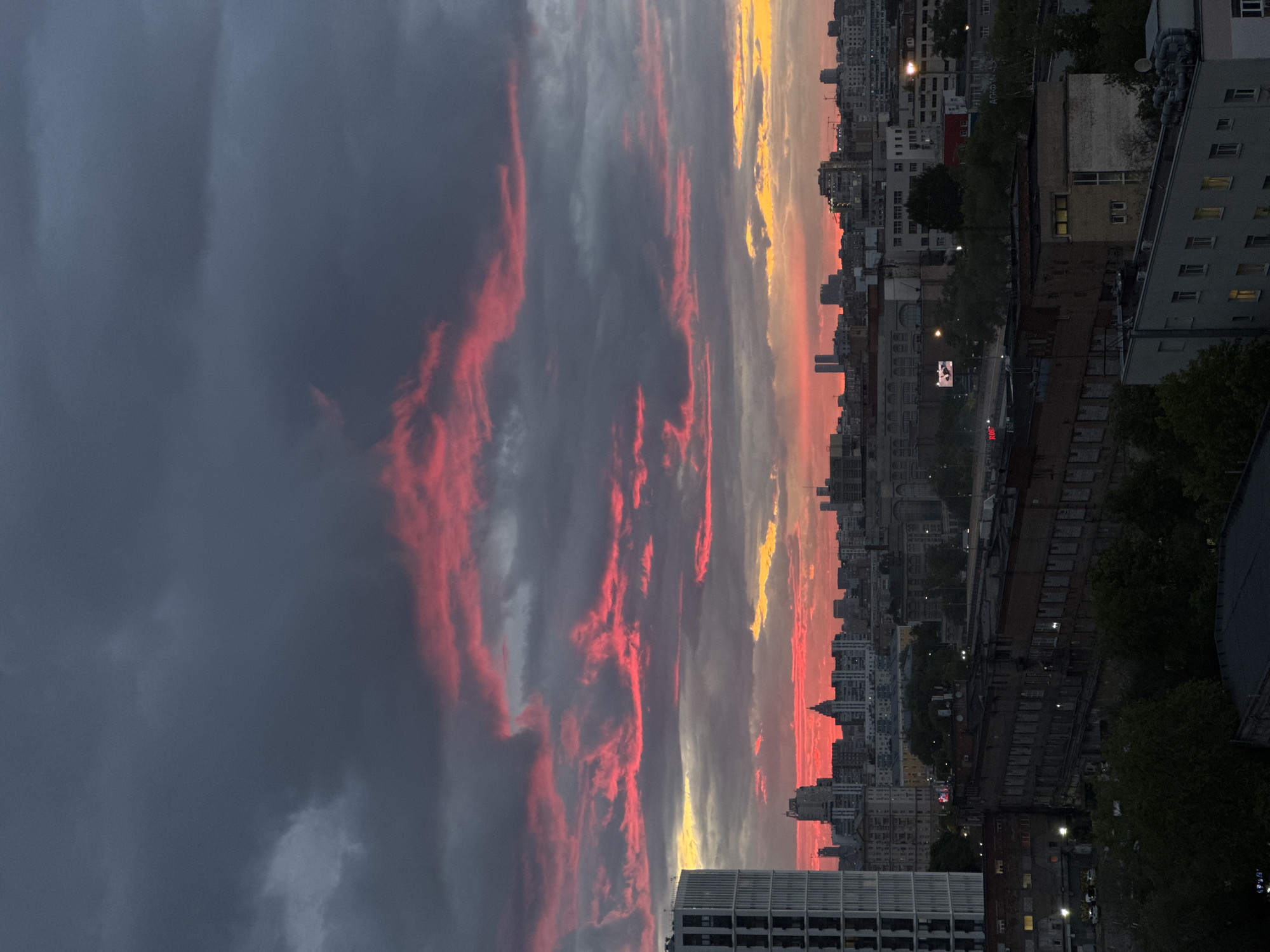 The image shows a view of Moscow, Russia, at sunset. The sky is filled with clouds ranging in color from dark gray to vibrant red and yellow. Buildings of varying heights and architectural styles stretch across the horizon. Some buildings are illuminated by streetlights and interior lights. Trees are scattered throughout the cityscape.