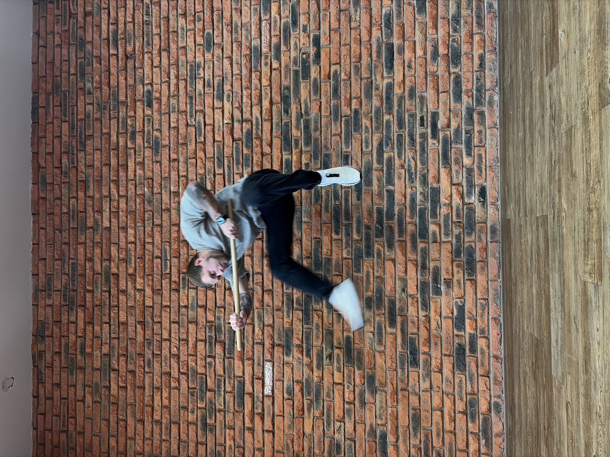 A full-length, high-angle shot captures a young man in an unusual, gravity-defying pose against a textured brick wall, giving the impression he is standing on a vertical surface. The image is oriented sideways, with the man appearing to walk or strike a dynamic pose on the brickwork.
The sole subject is a male with short hair and a beard, wearing a light gray long-sleeved shirt, dark trousers, and white athletic shoes. Tattoos are visible on his right forearm. He is holding a light-colored wooden stick or dowel horizontally with both hands, positioned across his body. His expression is neutral, possibly with a subtle smirk, and he appears focused on his action. His left foot is lifted and slightly blurred, suggesting movement or the culmination of a pose.
The setting consists primarily of a reddish-brown brick wall, which dominates the frame and acts as the "ground" in this inverted perspective. The bricks have dark, almost charred, markings, creating a rustic and distressed appearance. To the right of the brick wall, a strip of light brown wooden flooring runs vertically, creating a contrasting texture. On the far left, a plain, light gray wall is visible, with a small, irregular mark or hole near the bottom left corner. The lighting is bright and even, characteristic of an indoor environment. There is no visible text in the image.
This artistic photograph captures a moment of dynamic balance and playful illusion, set in an indoor location in Chaikovsky, Russia.