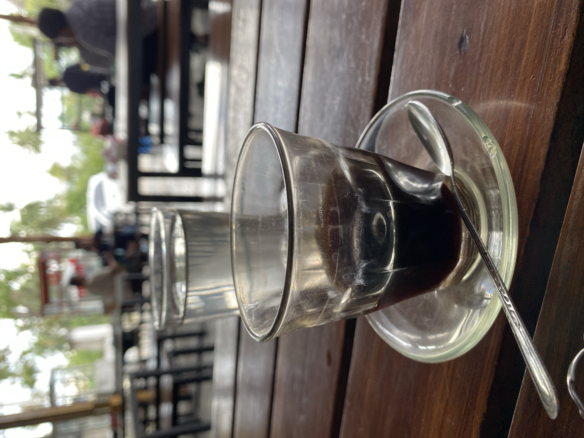 This is a close-up, eye-level shot of a glass of dark liquid, likely coffee, on a saucer with a spoon, placed on a wooden table. The glass is partially filled, and the liquid has a rich, dark color. The saucer is clear glass, and the spoon is silver and appears to have a decorative pattern. In the background, a second glass of clear liquid is visible, as well as blurred figures of people and tables, suggesting an outdoor or semi-outdoor seating area of a cafe or restaurant. The environment appears to be in Cotgadong, Indonesia, indicated by the tropical foliage visible in the blurred background and the general ambiance of a relaxed outdoor dining establishment. The overall lighting is diffused, suggesting an overcast day or shaded area.