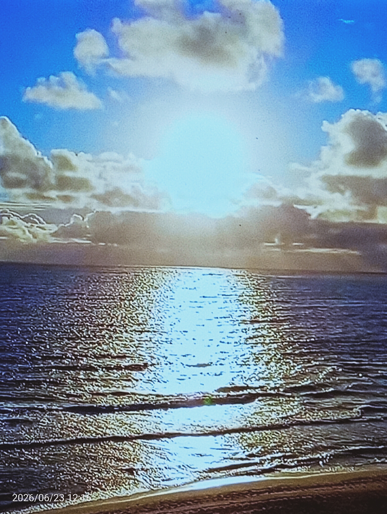 The image displays a vast body of water meeting a beach under a bright sky filled with clouds. The sun is high in the sky, with its rays reflecting off the water, creating a shimmering effect. The sky is a vibrant blue, interspersed with fluffy white and gray clouds. The water appears to be calm, with gentle waves lapping at the shore. A strip of sand forms the beach in the foreground. A timestamp in the bottom left corner of the image reads "2026/06/23 12:16". There are no people or specific objects of note visible in the scene. The location is listed as Unknown, Unknown.