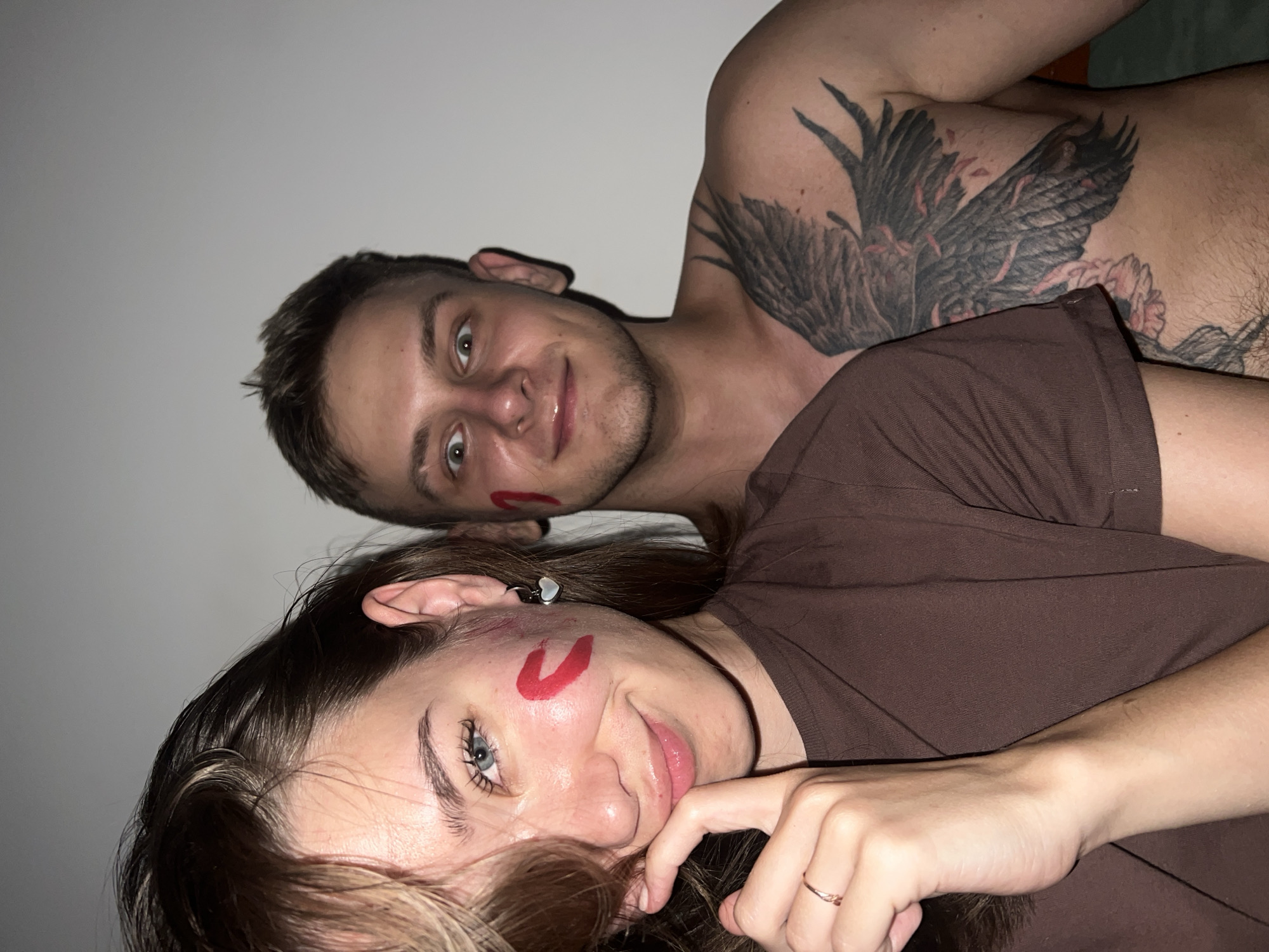 Two people are lying down close to each other, smiling at the camera. A man with short brown hair and a red mark on his face is behind a woman with long brown hair, blue eyes, and a similar red mark on her face. The man has a large tattoo of a bird on his chest. The woman is wearing a brown t-shirt and a small earring. The background is a plain white wall. The location is not evident from the photo. There is no information that suggests the location is Nam Natural Heritage Area Trang, Vietnam.