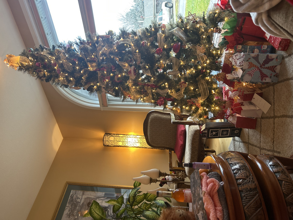 A decorated Christmas tree stands by a window, surrounded by wrapped gifts in various colors and patterns. The tree is adorned with lights, red ornaments, and gold ribbons. A vintage armchair with a red pillow sits nearby. A floor lamp provides ambient lighting. In the foreground, a carved wooden table holds decorative items including candles, a plant, and a plush animal. A framed picture hangs on the wall.