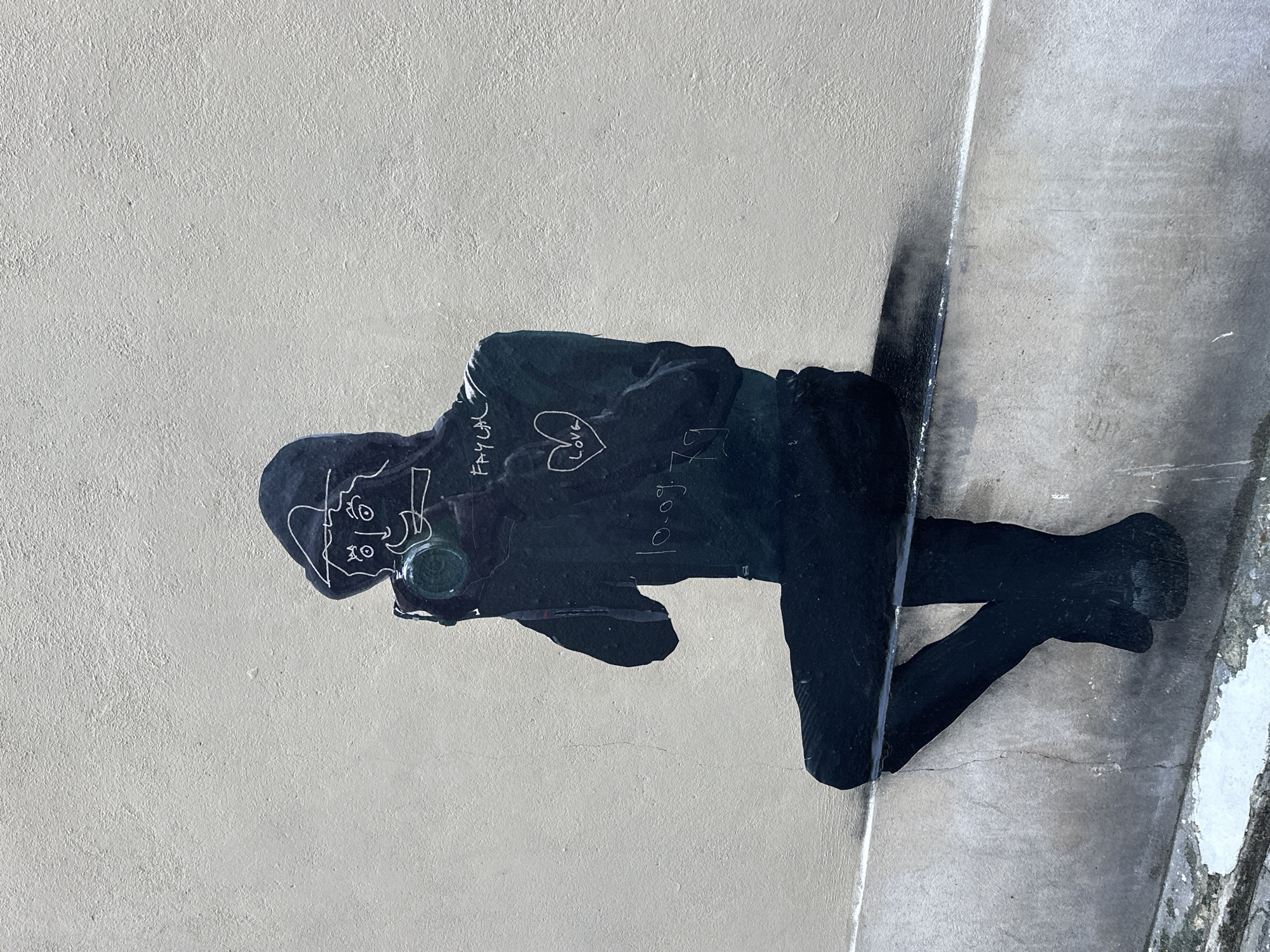 An outdoor image captures a piece of street art on a light-colored, textured building wall in Paris, France. The artwork depicts a stylized, full-body silhouette of a person rendered primarily in black, sitting on a dark horizontal ledge that protrudes from the wall. The figure faces toward the viewer and slightly to the right, holding a camera with both hands to their eye, as if taking a photograph. The camera lens features a distinct green element. White lines define the figure's face, showing circular eyes, a nose, a small mouth, and a peaked cap or hood. Additional white outlines detail the hands and various elements on the black clothing, which appears to be a hooded jacket and trousers. Inscribed on the chest area of the jacket are the words "FAYCAL," a heart symbol containing the word "LOVE," and the numbers "10.09.79." Below the dark ledge, the wall transitions to a rougher, darker gray surface with visible wear and discoloration, suggesting a lower architectural feature or ground level. The overall scene is static, focusing entirely on the detailed mural against the urban backdrop.